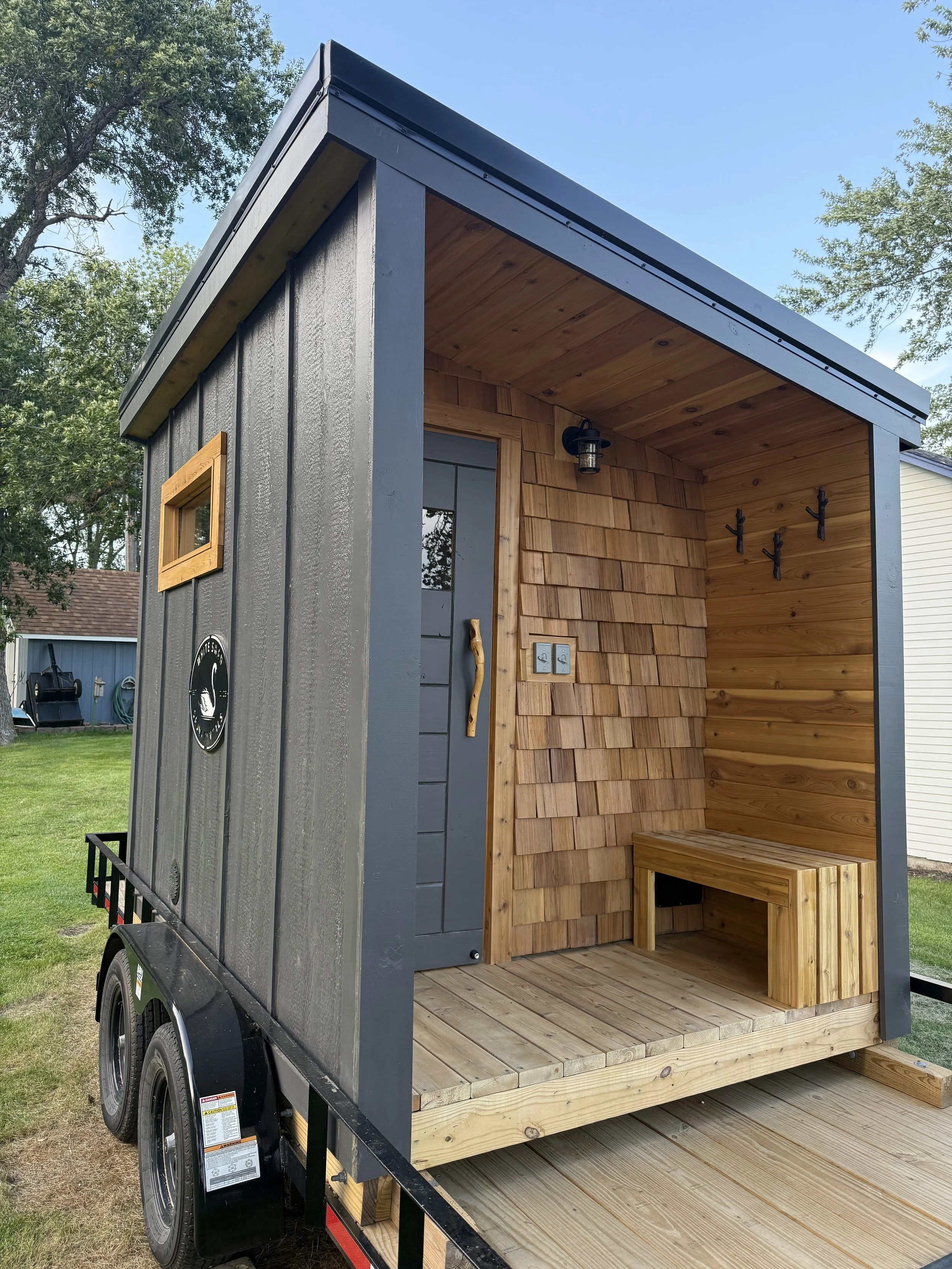 Outdoor sauna with a dark gray exterior, wooden interior, a bench, coat hooks, and an exterior light.