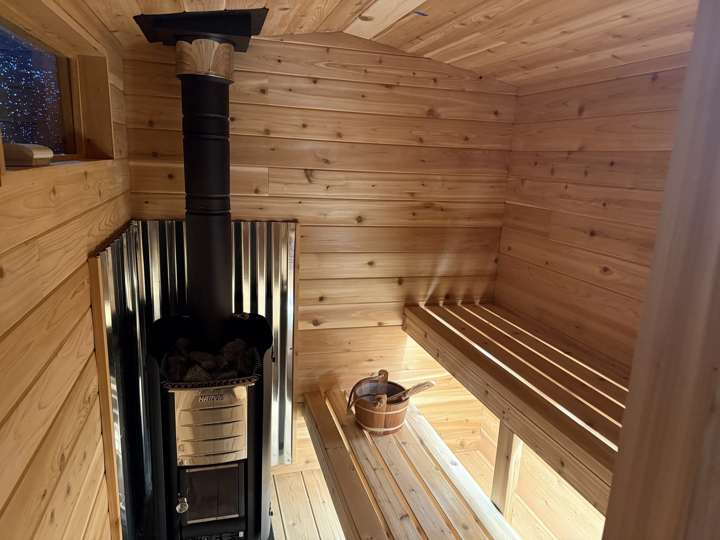 Interior of a small wooden sauna with a wood bench, a bucket with a wooden ladle, and a stove with stones. The walls and ceiling are made of cedar wood.