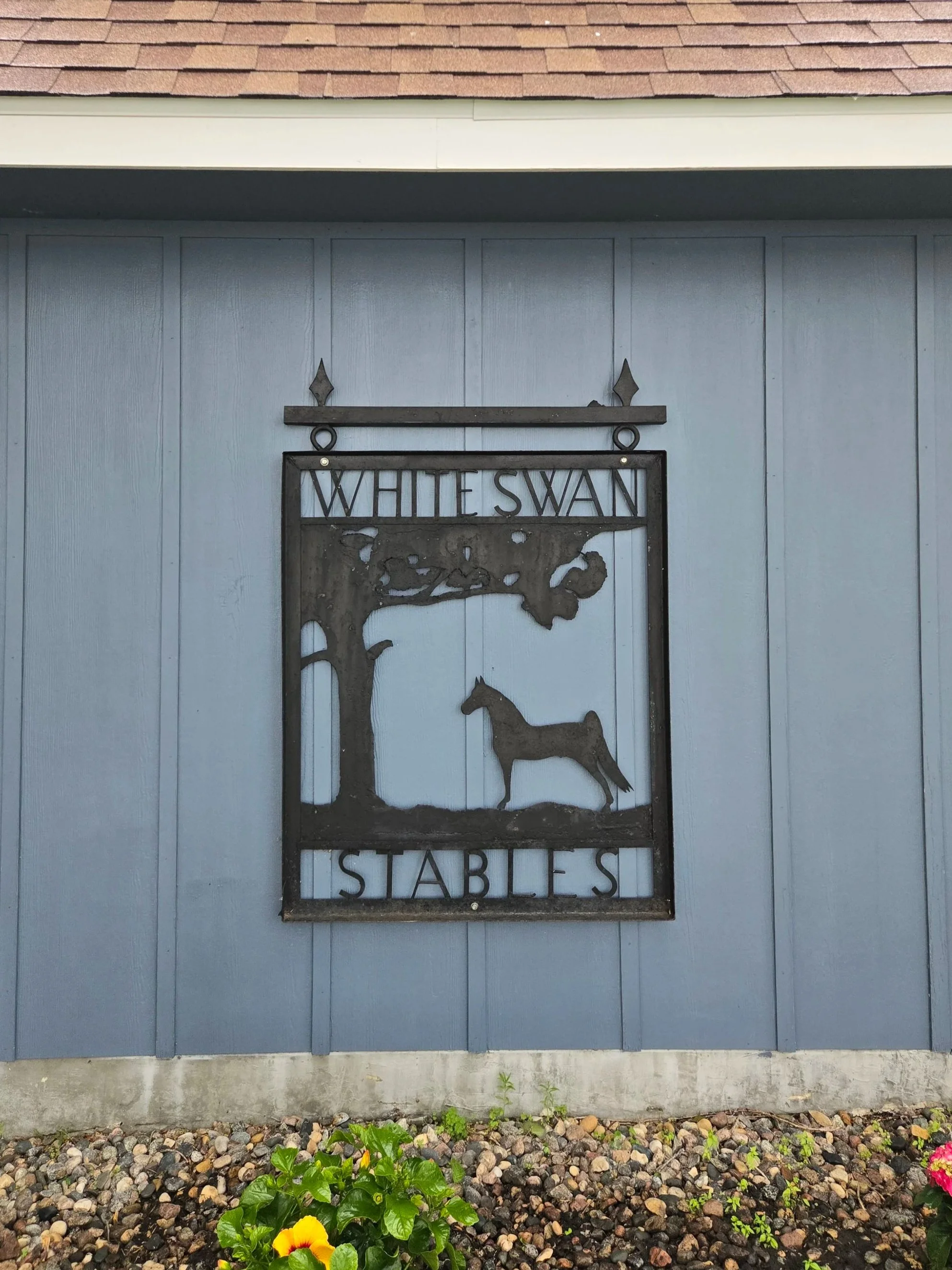 A metal sign reading 'White Swan Stables' on a gray wall, with a silhouette of a horse under a tree, located above some flowers and small plants.