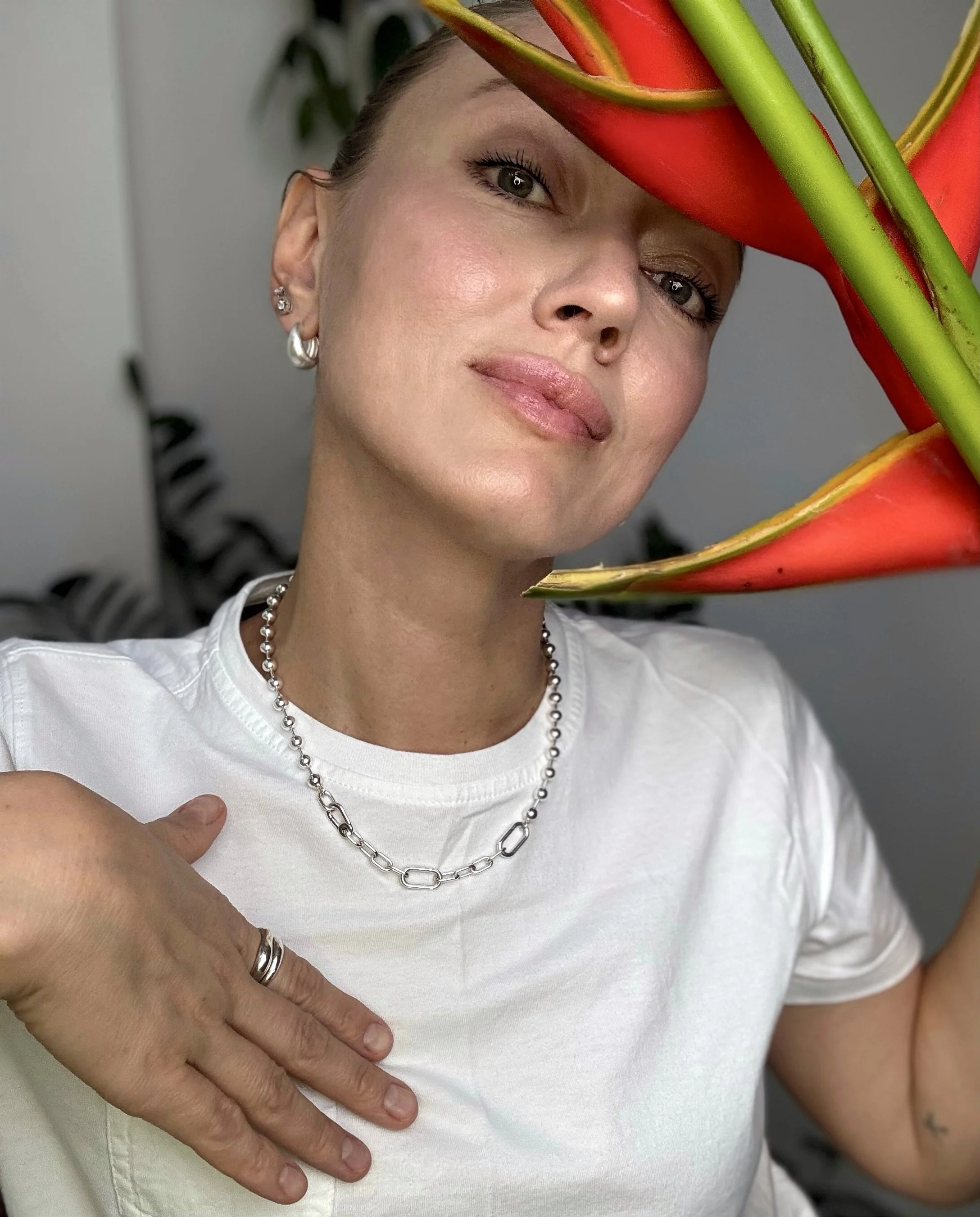 A woman with earrings and a necklace, wearing a white shirt, holding her chest with her left hand.