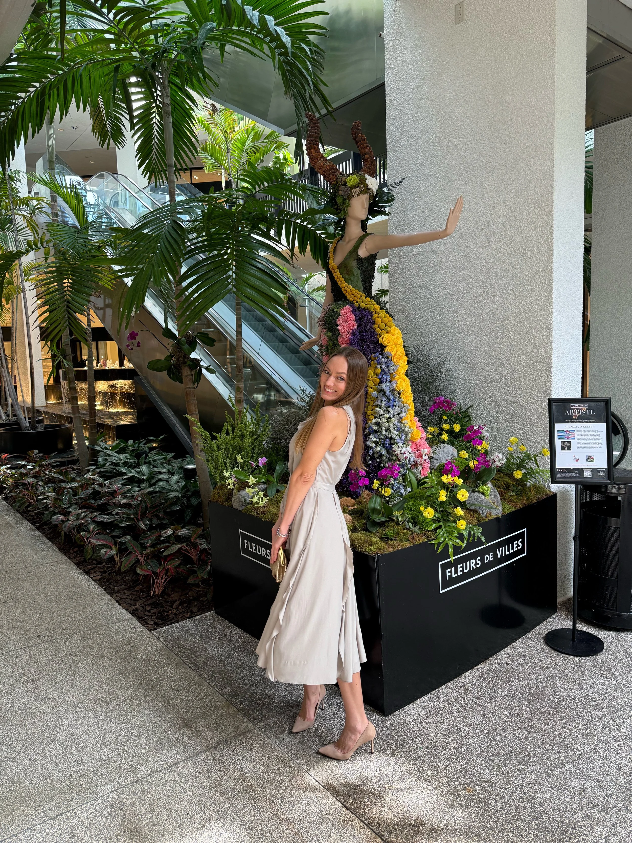A woman in an elegant beige dress and high heels smiling and posing next to a floral display featuring a mannequin with a horned headdress made of flowers and greenery, in a shopping mall decorated with various green plants and flower arrangements.