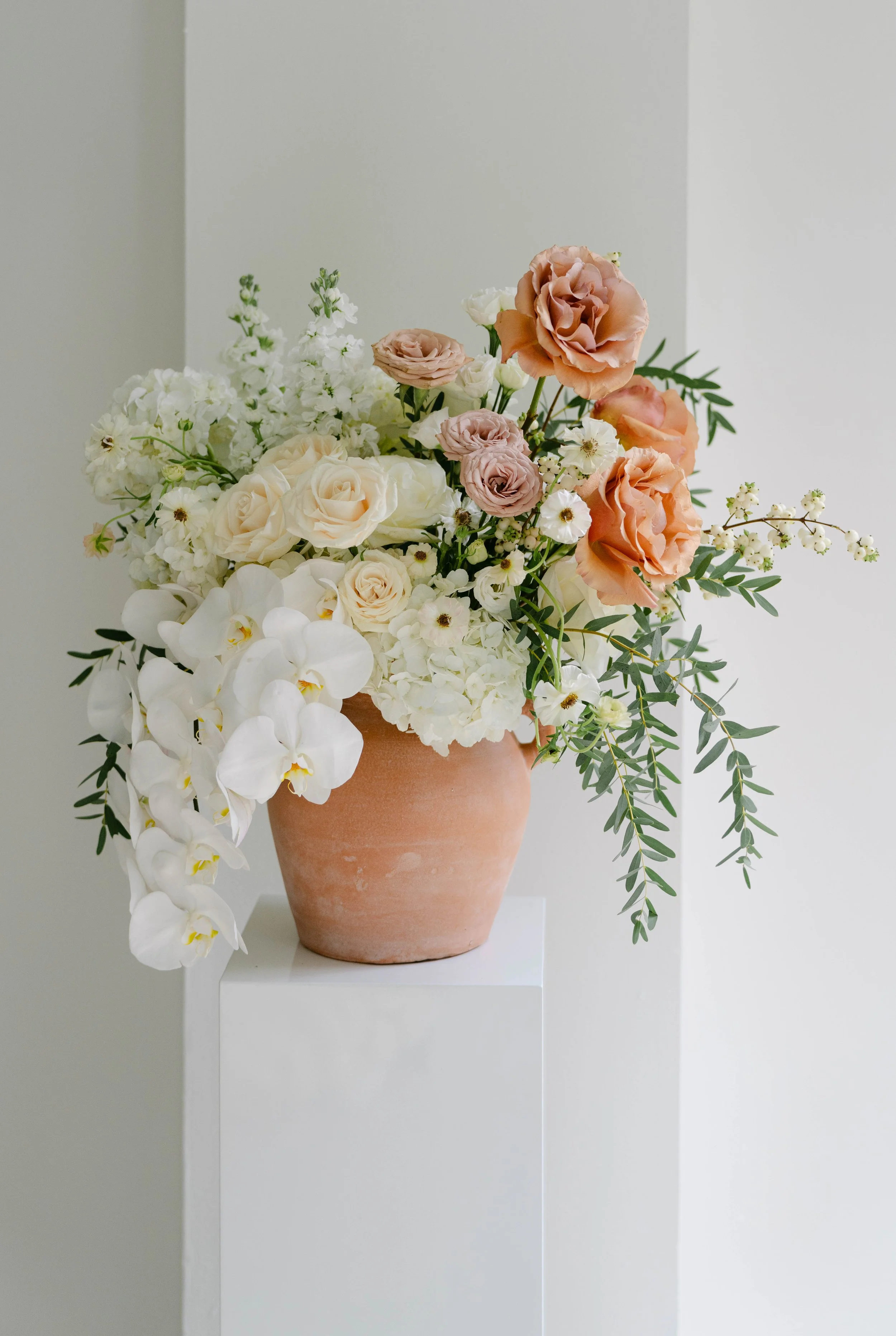 White orchids, cream and peach roses with trailing greenery in a terracotta vase by KateAsFlowers.