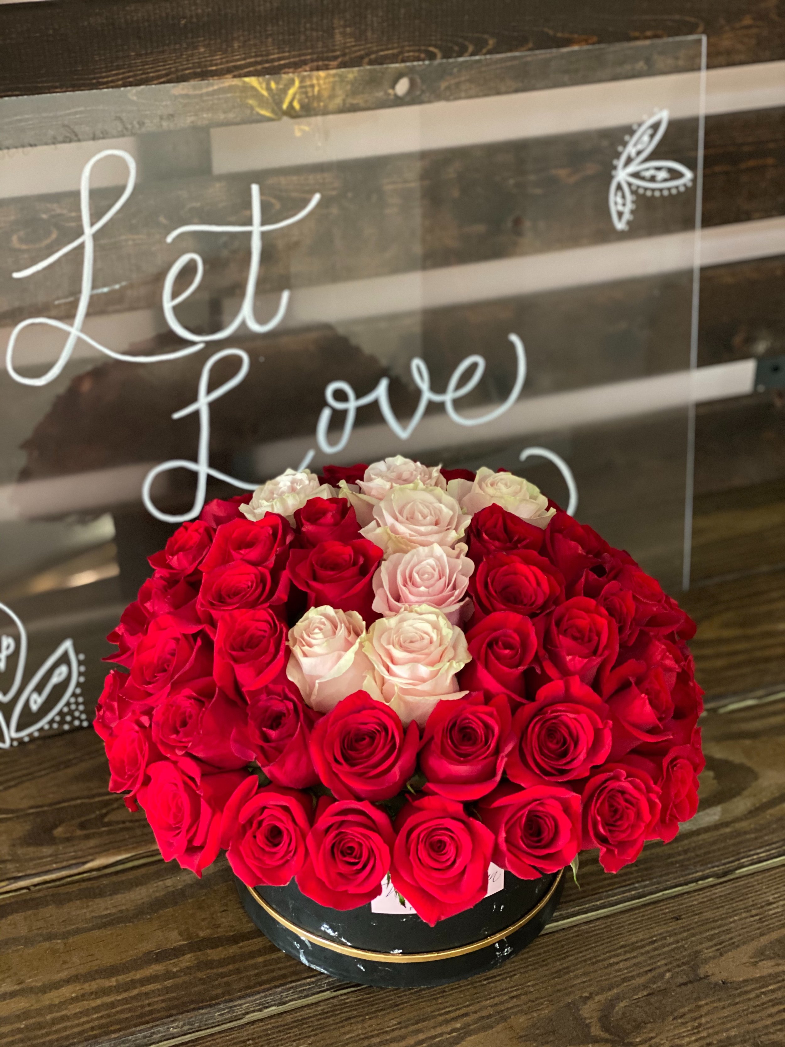 A round bouquet of red and cream roses in a black vase with a gold rim, placed on a wooden surface. In the background, there is a glass sign with white lettering that says 'Let Love' and decorated with a small floral design.