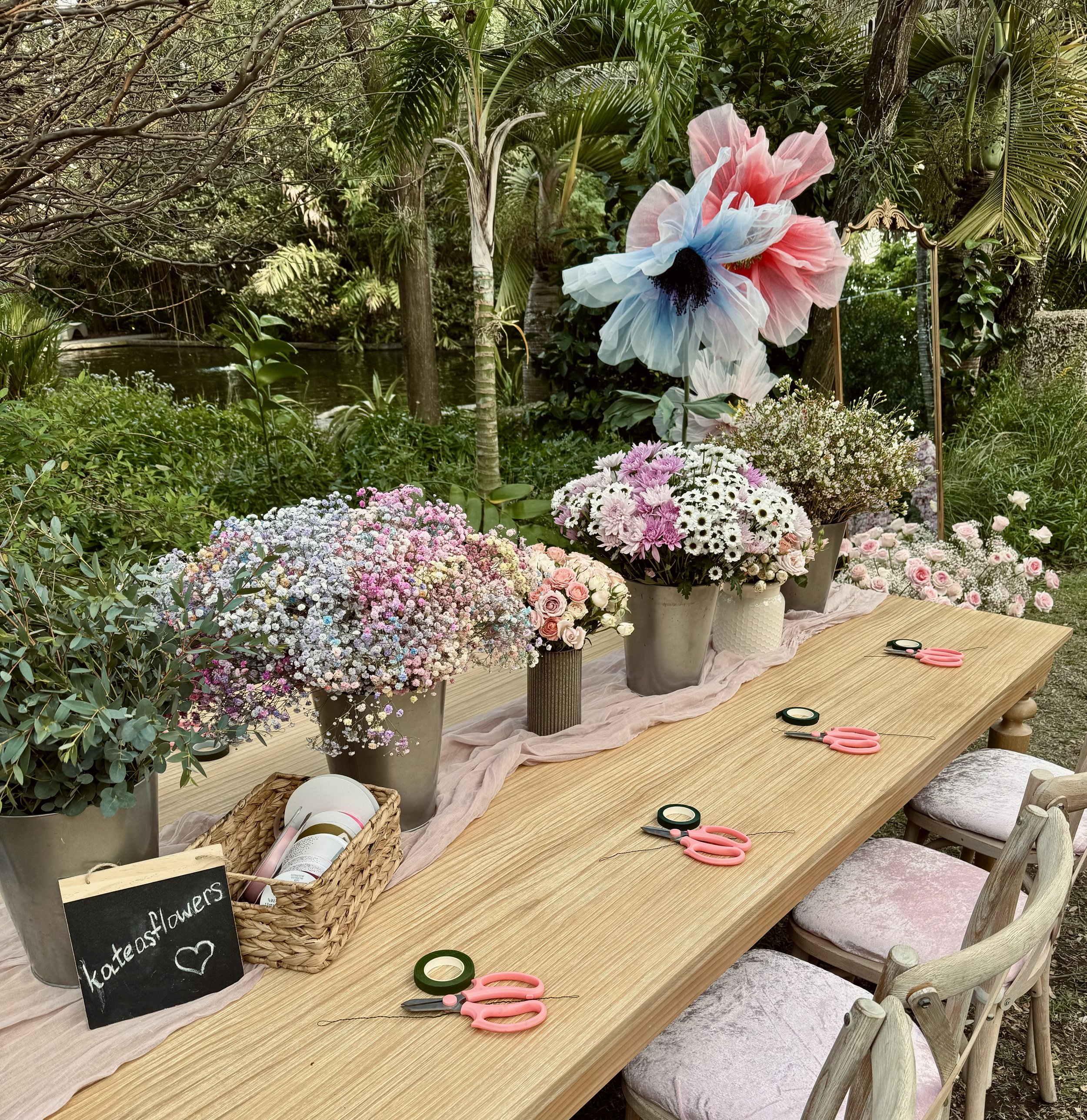 A wooden table decorated with various flower arrangements and gardening tools, set outdoors with lush greenery and a small pond in the background.