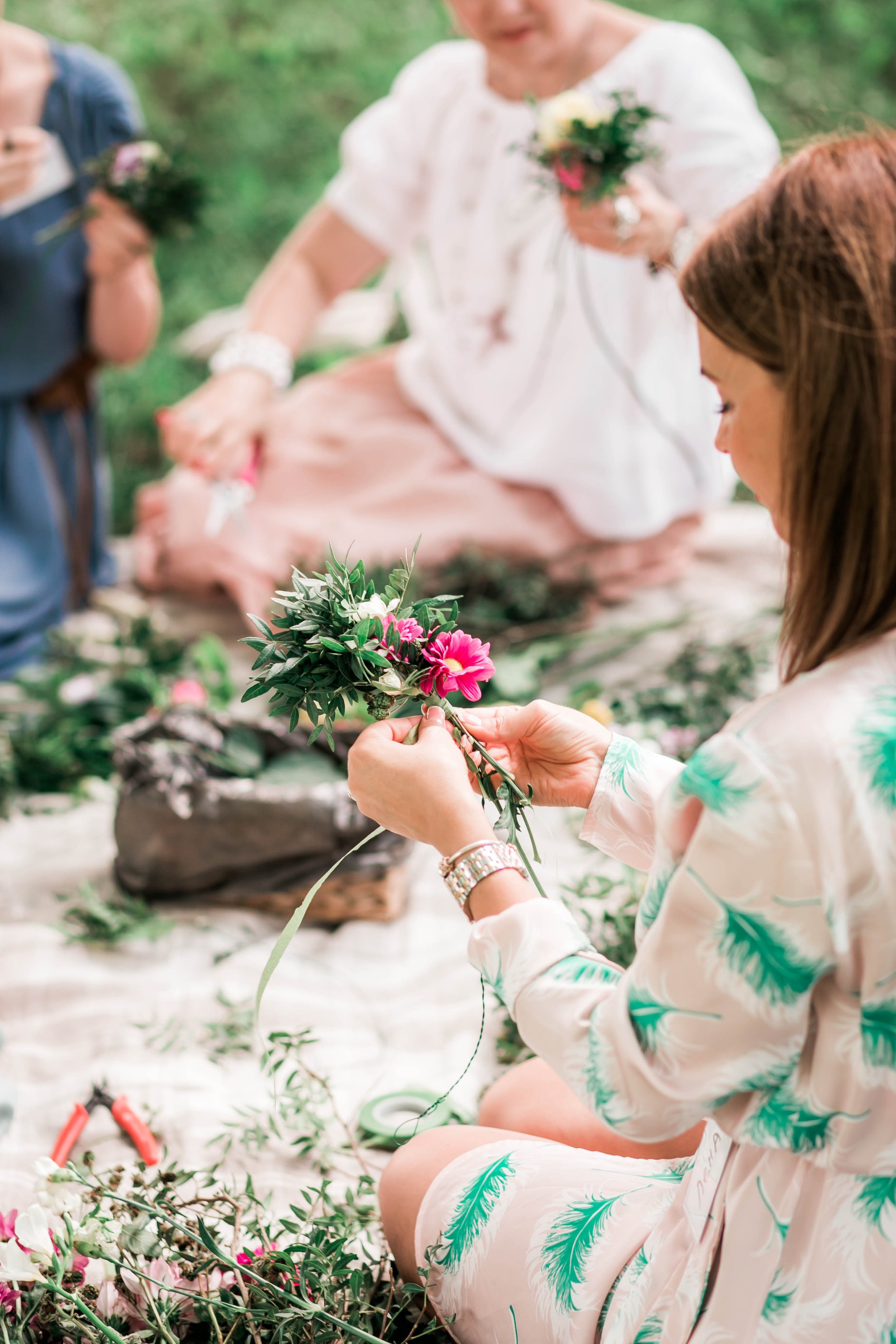 A group of women sitting on a white blanket outdoors, making floral arrangements with pink and white flowers and greenery.