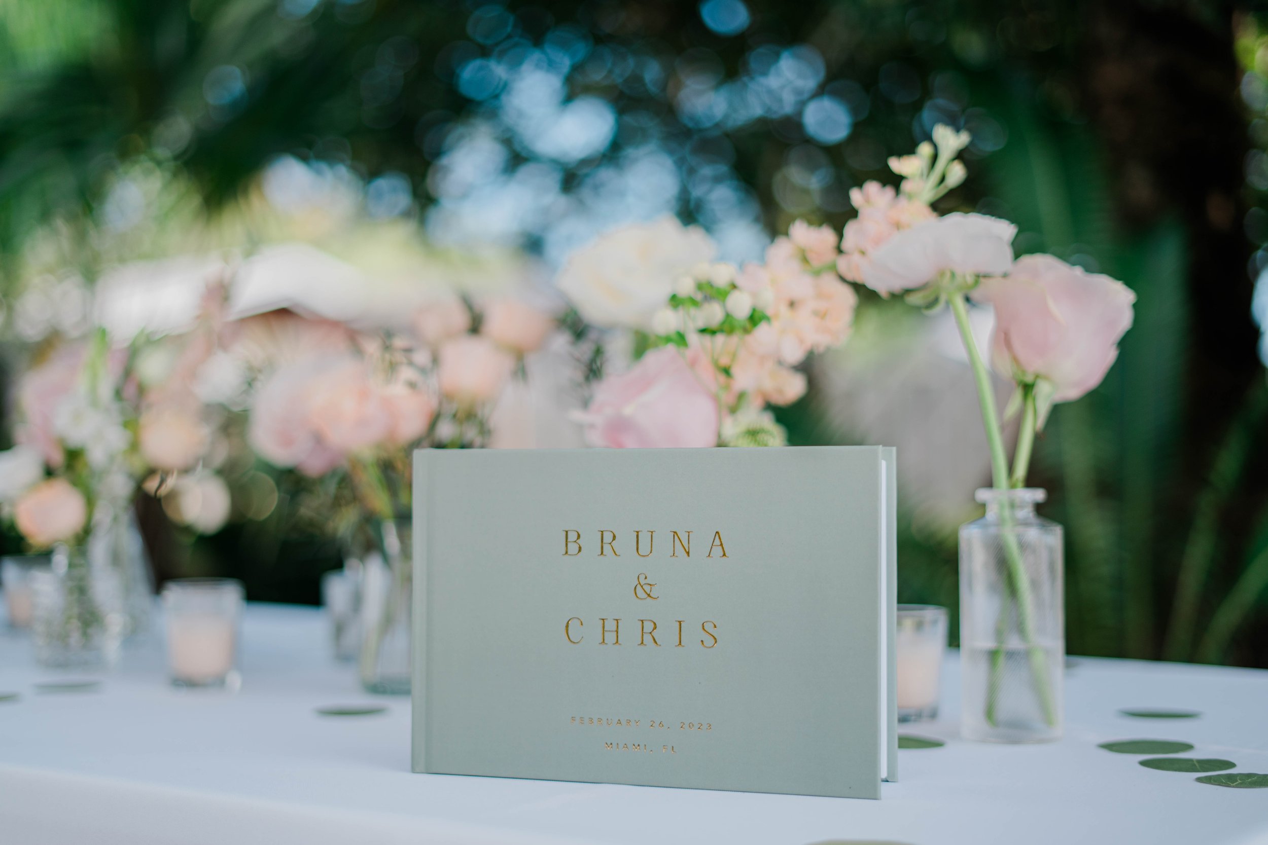 A wedding table sign with the names Bruna and Chris, dated February 26, 2023, in Miami, Florida, surrounded by pink and white flowers in glass vases.