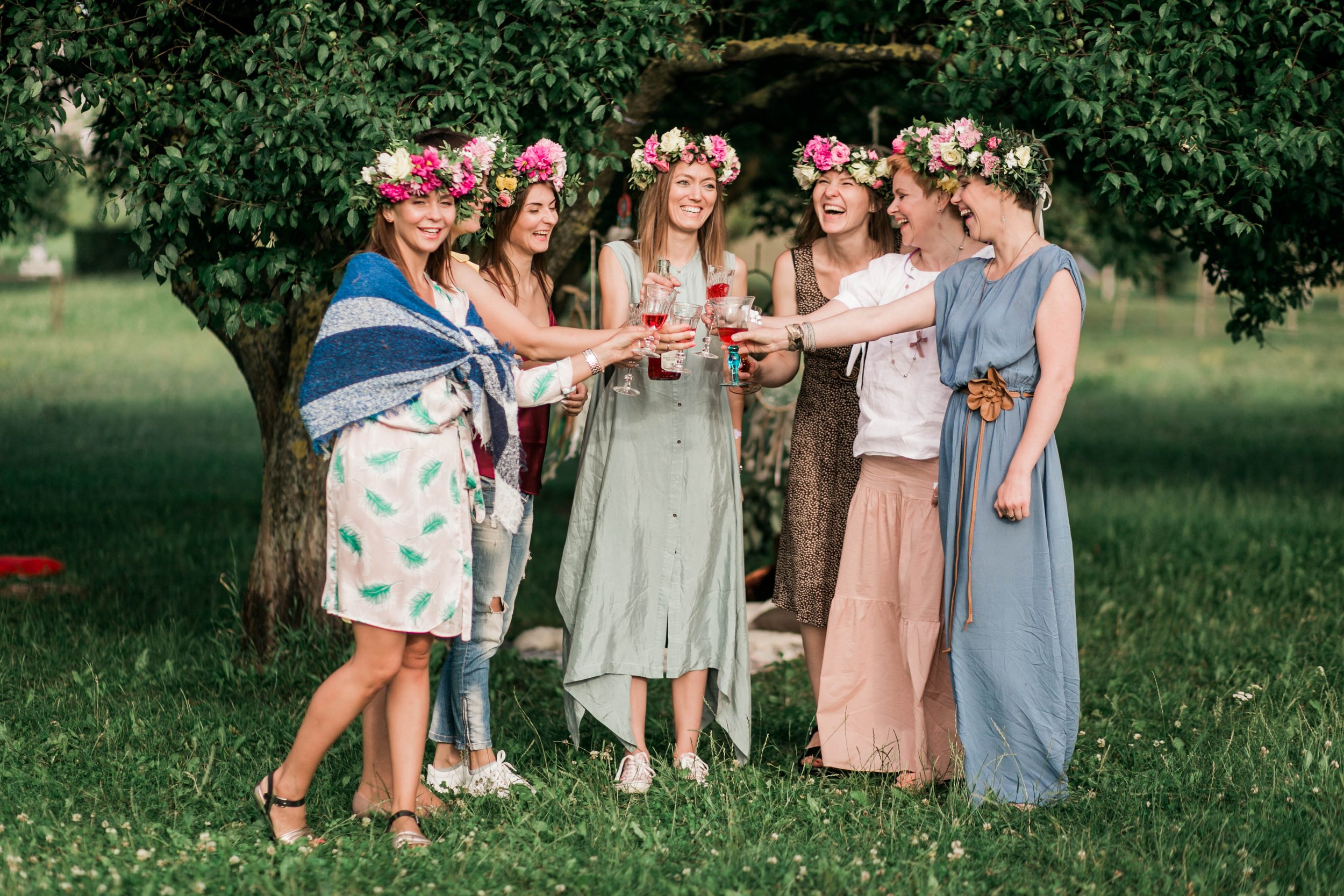 Group of women wearing floral crowns toasting with drinks outdoors, standing under a tree.