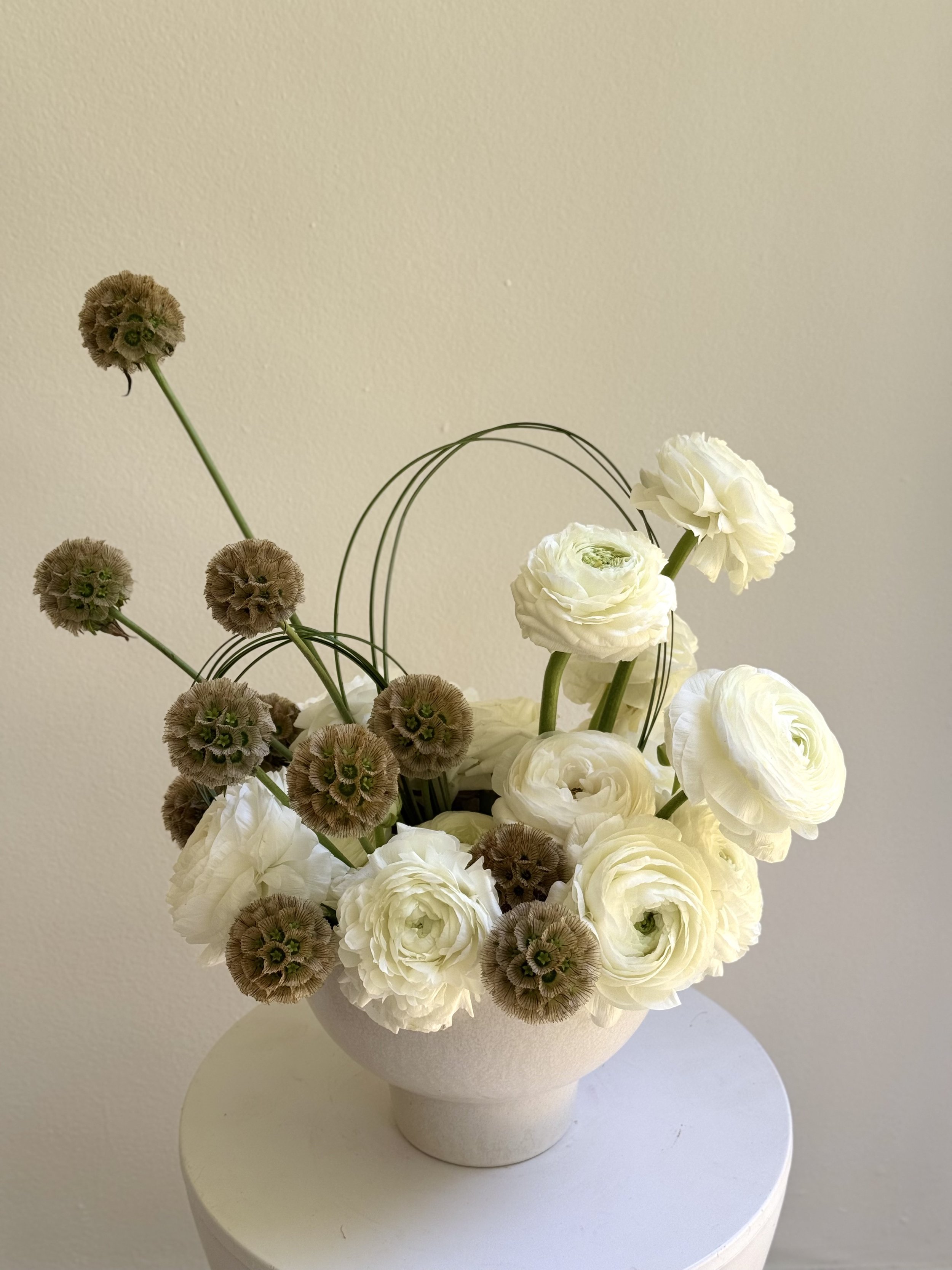 A white ceramic vase filled with white ranunculus flowers and brown seed heads, arranged on a round white table against a plain beige wall.