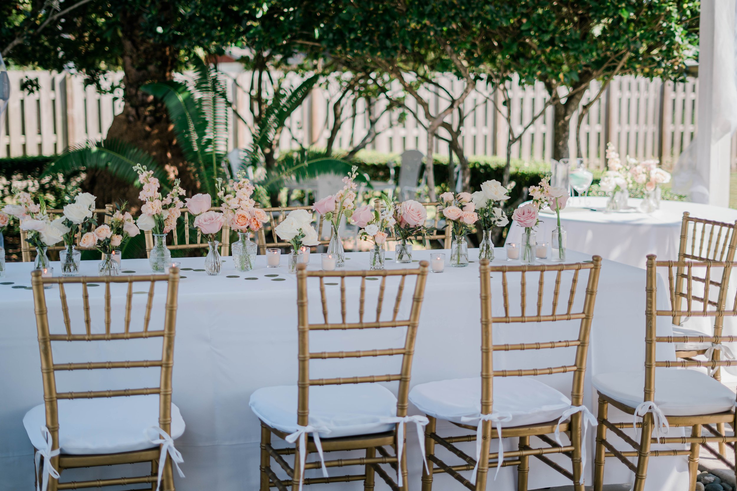 Outdoor dining table decorated with pink and white flowers in glass vases, surrounded by gold chairs with white cushions, set for a celebration outdoors under trees with a white fence in the background.