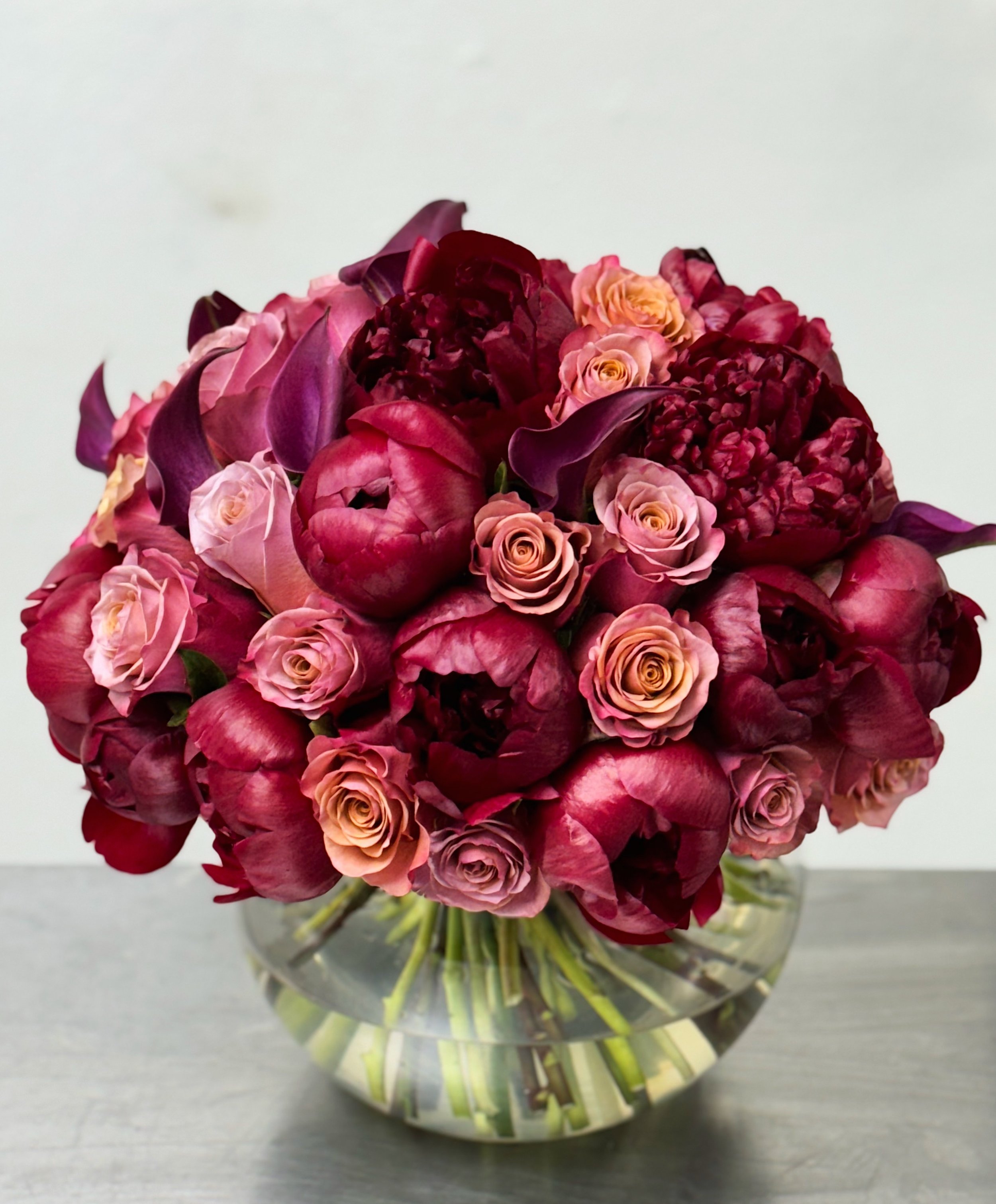 A bouquet of deep burgundy and pink flowers in a clear round glass vase on a gray surface.