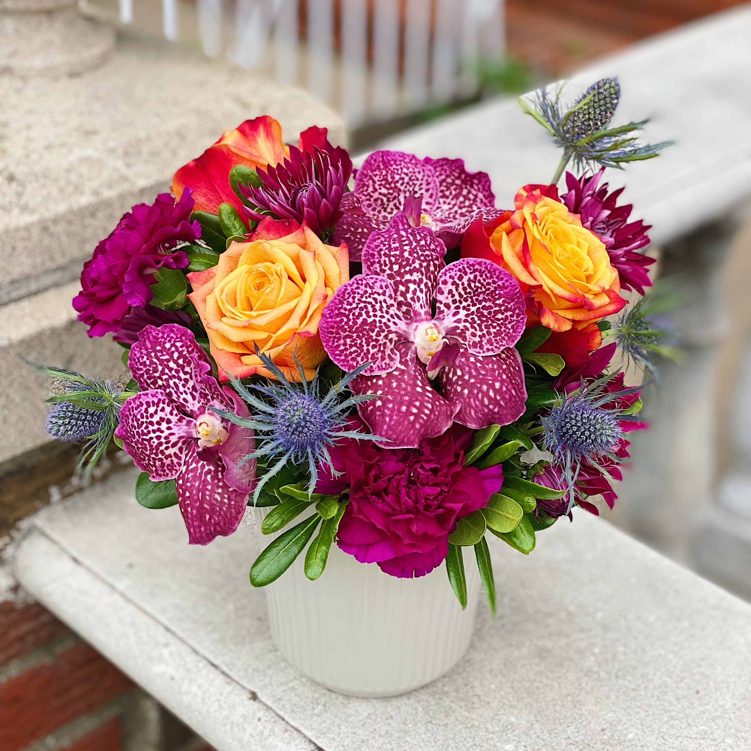 A colorful bouquet of various flowers, including yellow roses, purple orchids, pink carnations, and blue thistles, in a white ribbed vase on a light gray surface. Background includes a brick wall and a wooden railing.