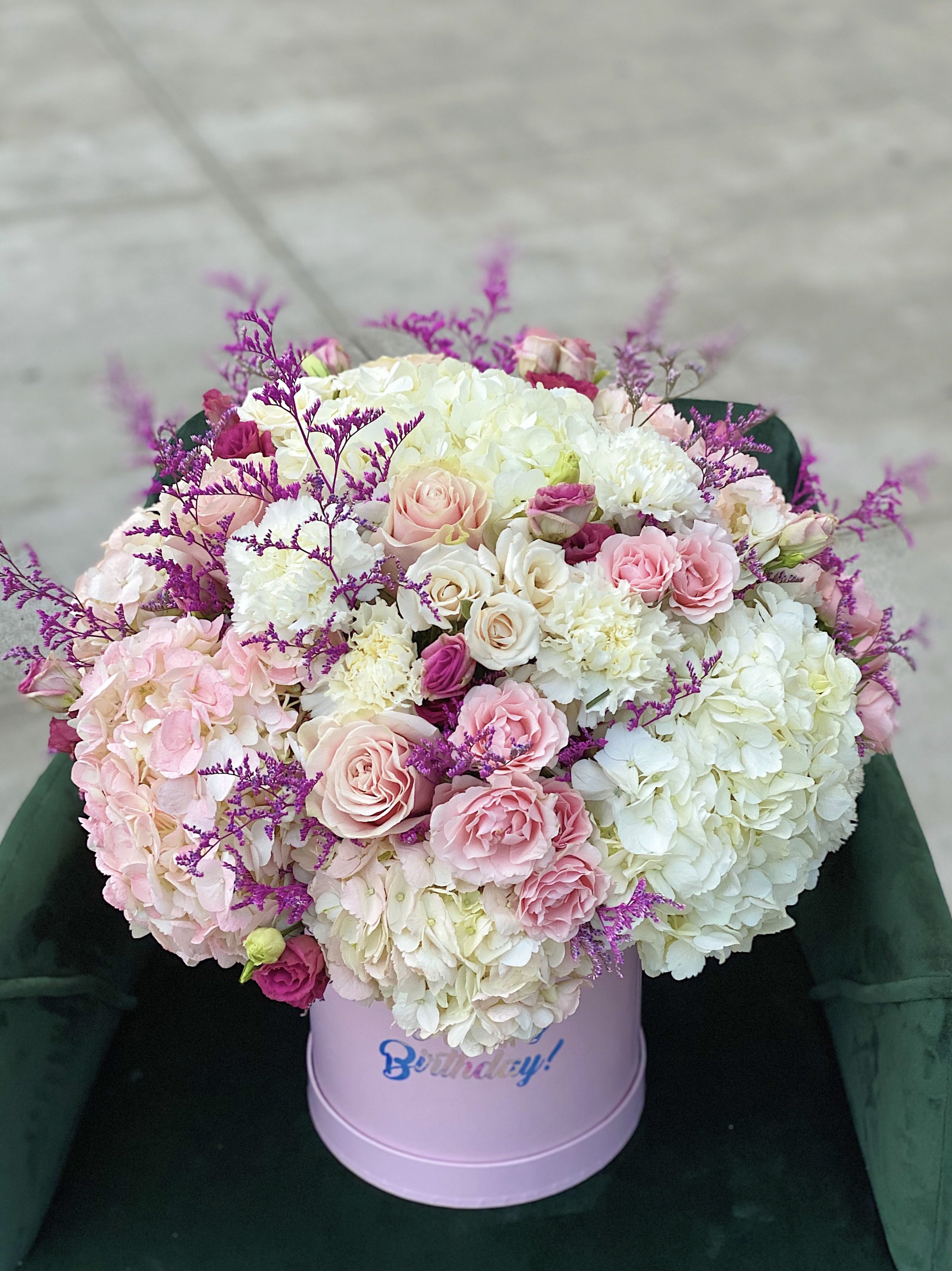 A pink birthday flower arrangement with hydrangeas, roses, carnations, and purple accent flowers in a pink hatbox with 'Birthday!' written on it.