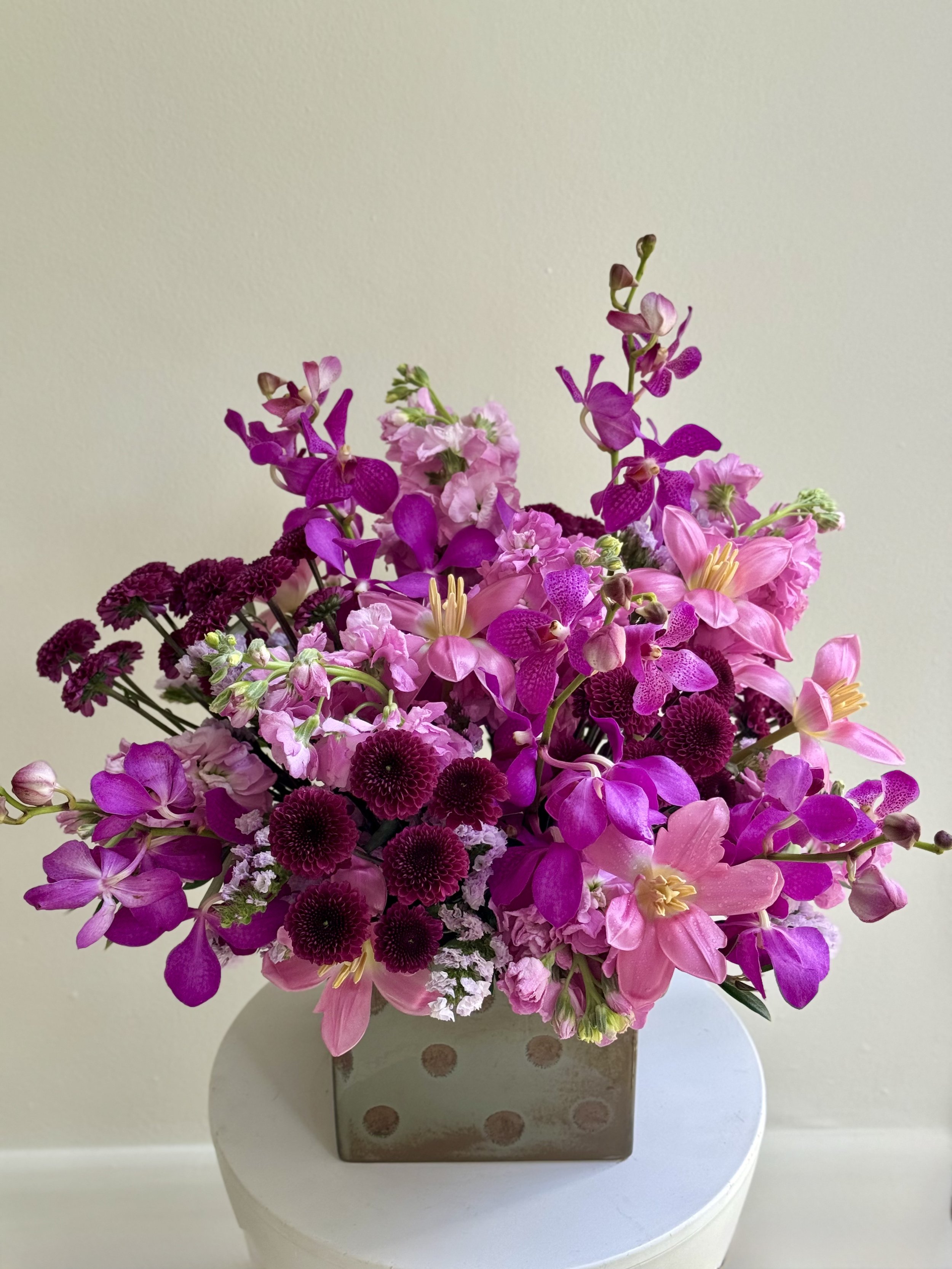 A bouquet of pink and purple flowers in a square metallic vase on a white round table against a plain beige wall.