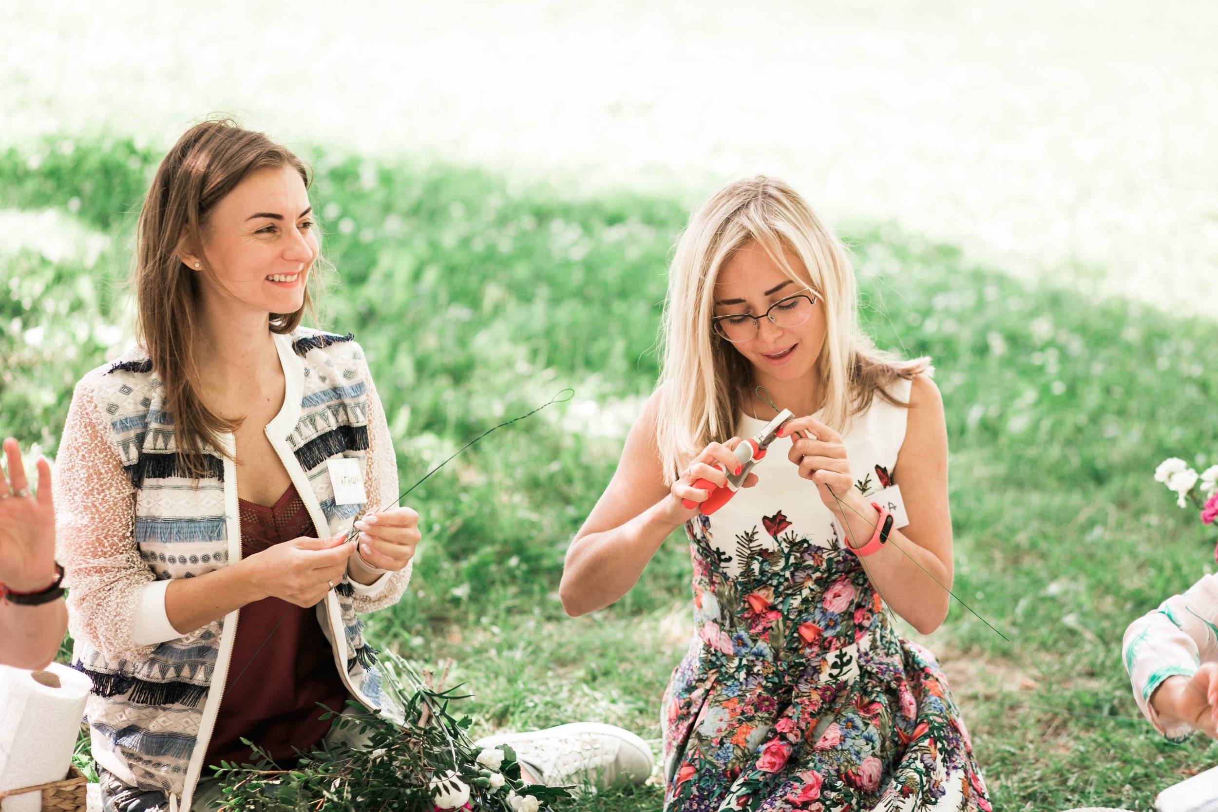 Two women sitting on grass outdoors engaged in flower arranging, one is smiling and the other is using wire cutters, surrounded by flowers and greenery.