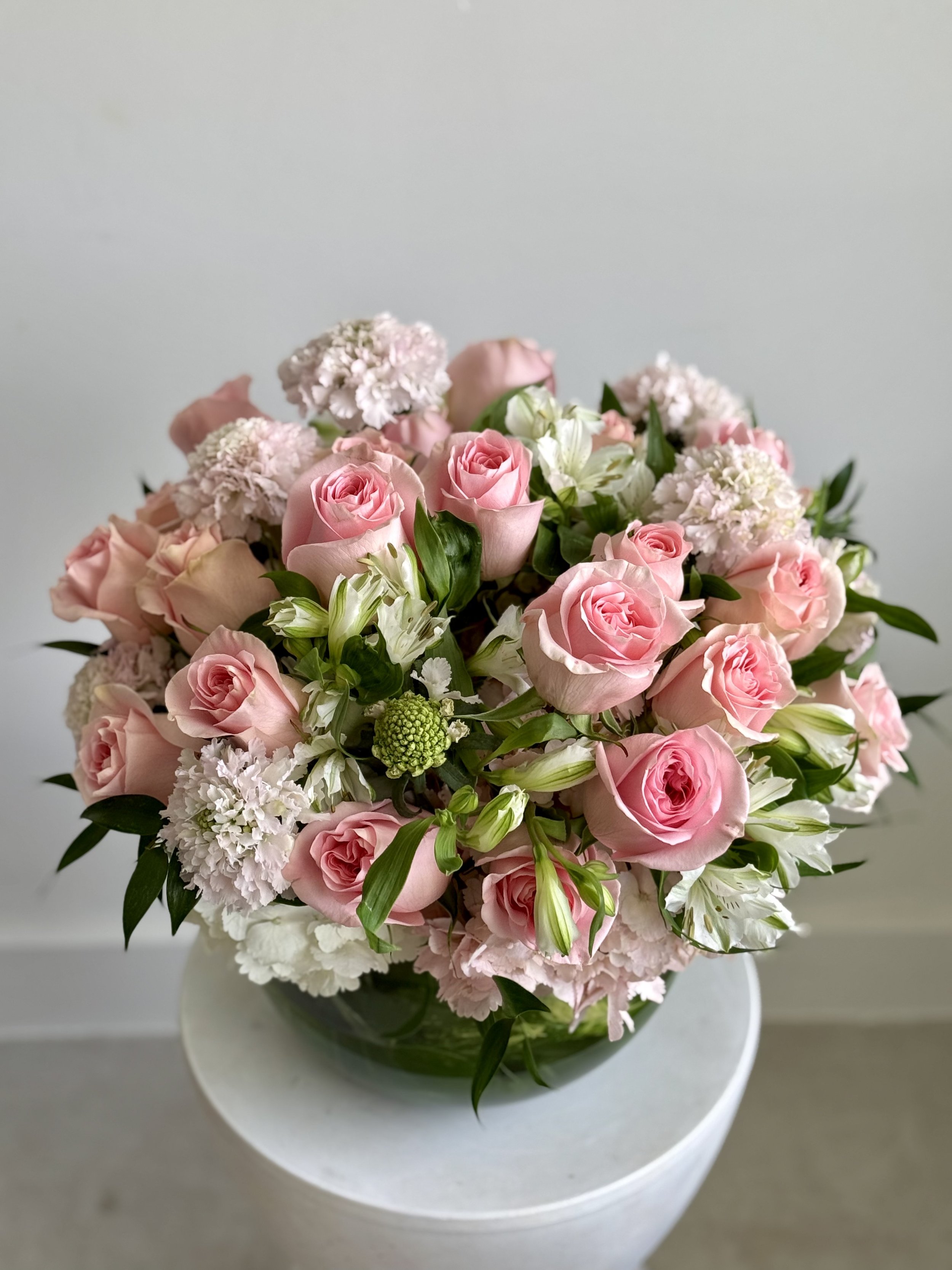 A bouquet of pink roses, white lilies, and white stock flowers in a glass vase on a white surface.