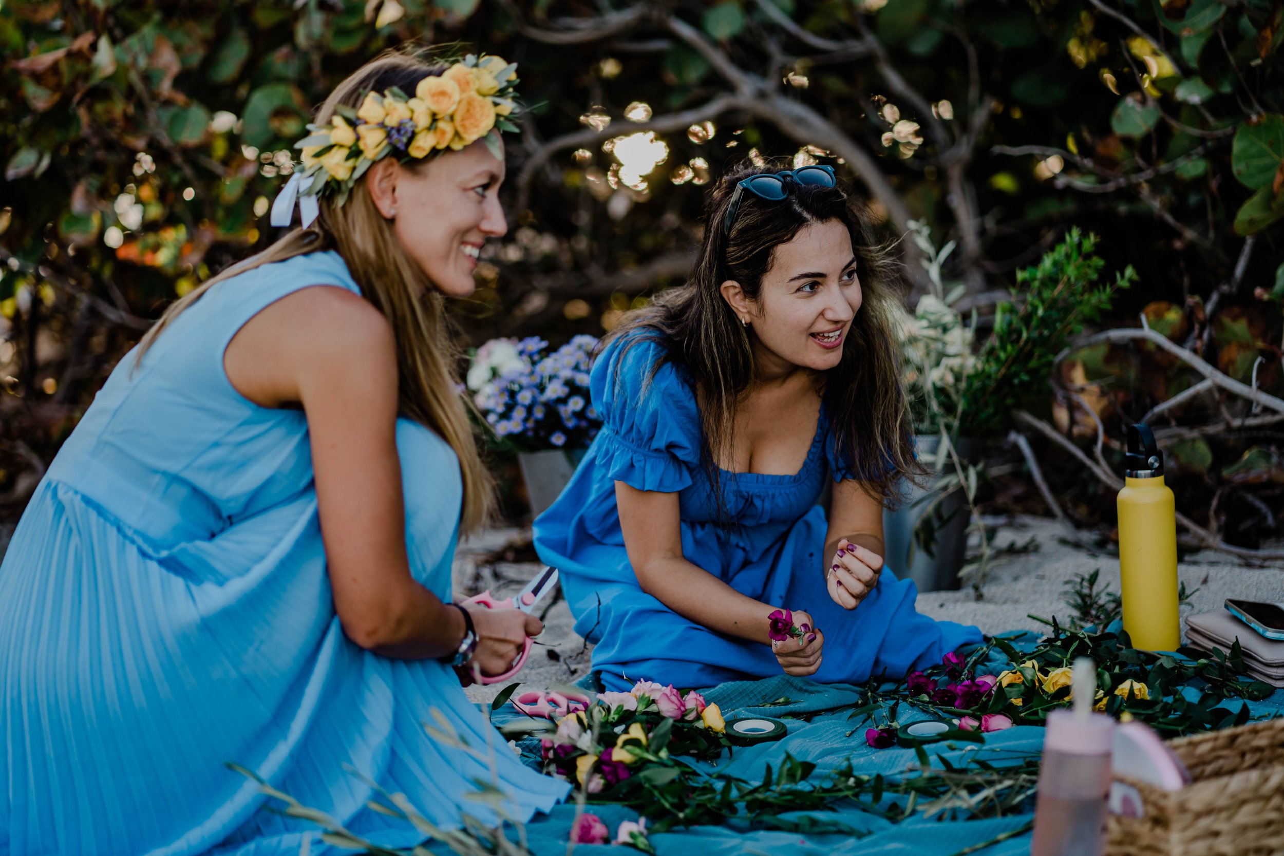 Two women sitting on a blanket in a natural outdoor setting, smiling and engaging in a flower-arranging activity, with flowers, scissors, and a water bottle nearby.