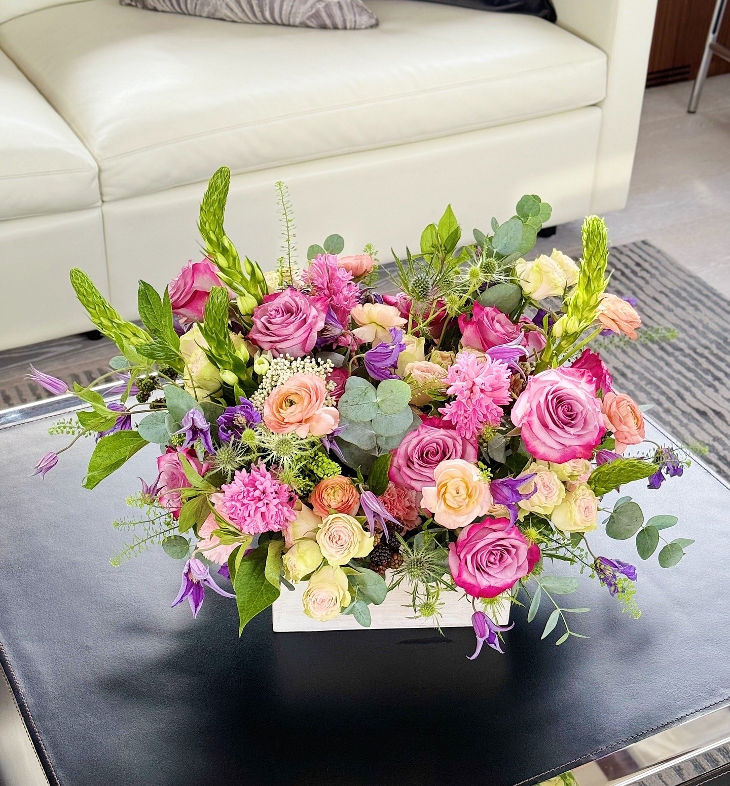Colorful floral arrangement with pink, purple, cream, and green flowers on a black leather table in a room with a white sofa and patterned rug.