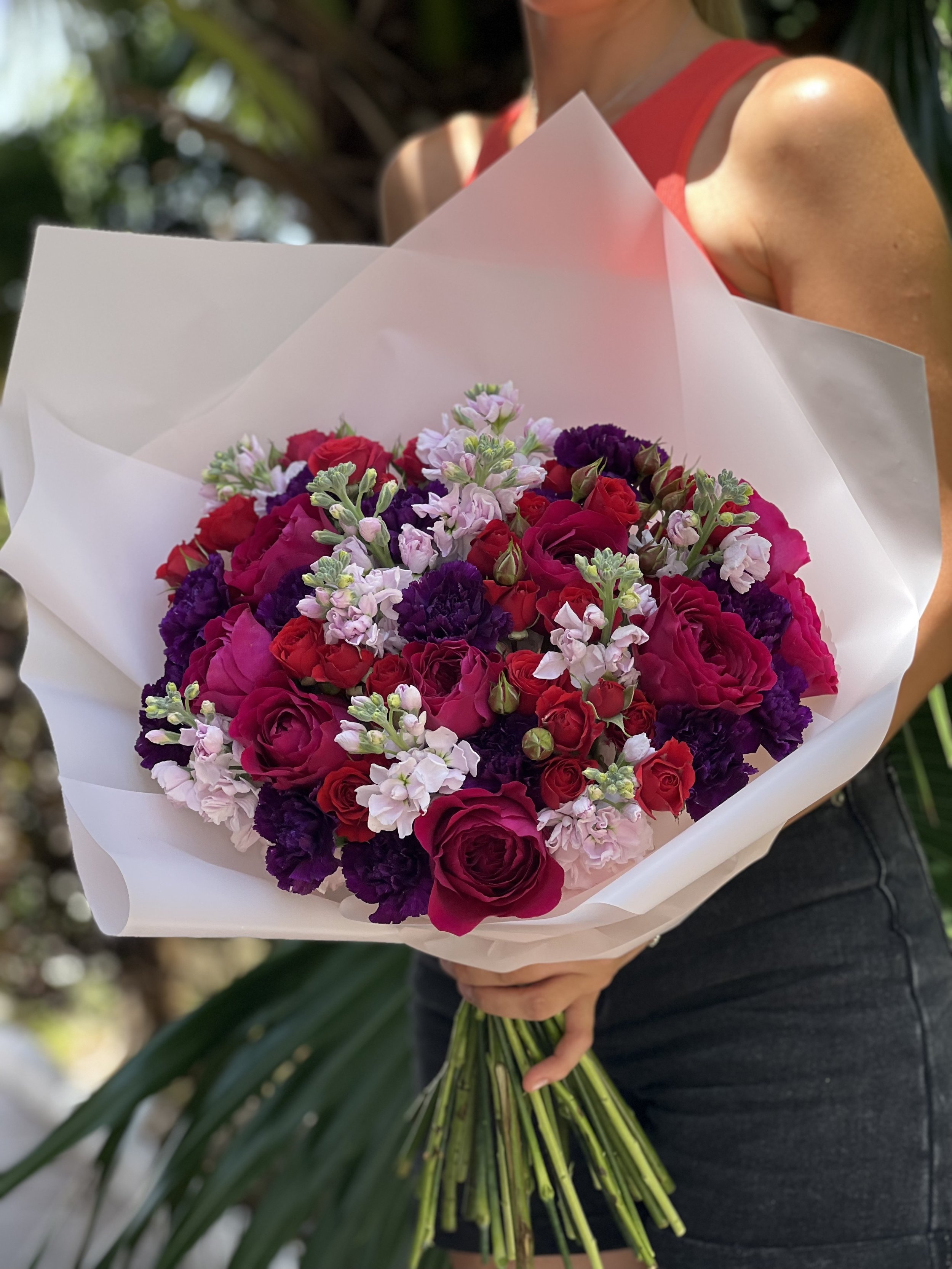 A woman wearing a red sleeveless top holding a large bouquet of pink, purple, and red flowers wrapped in white paper.