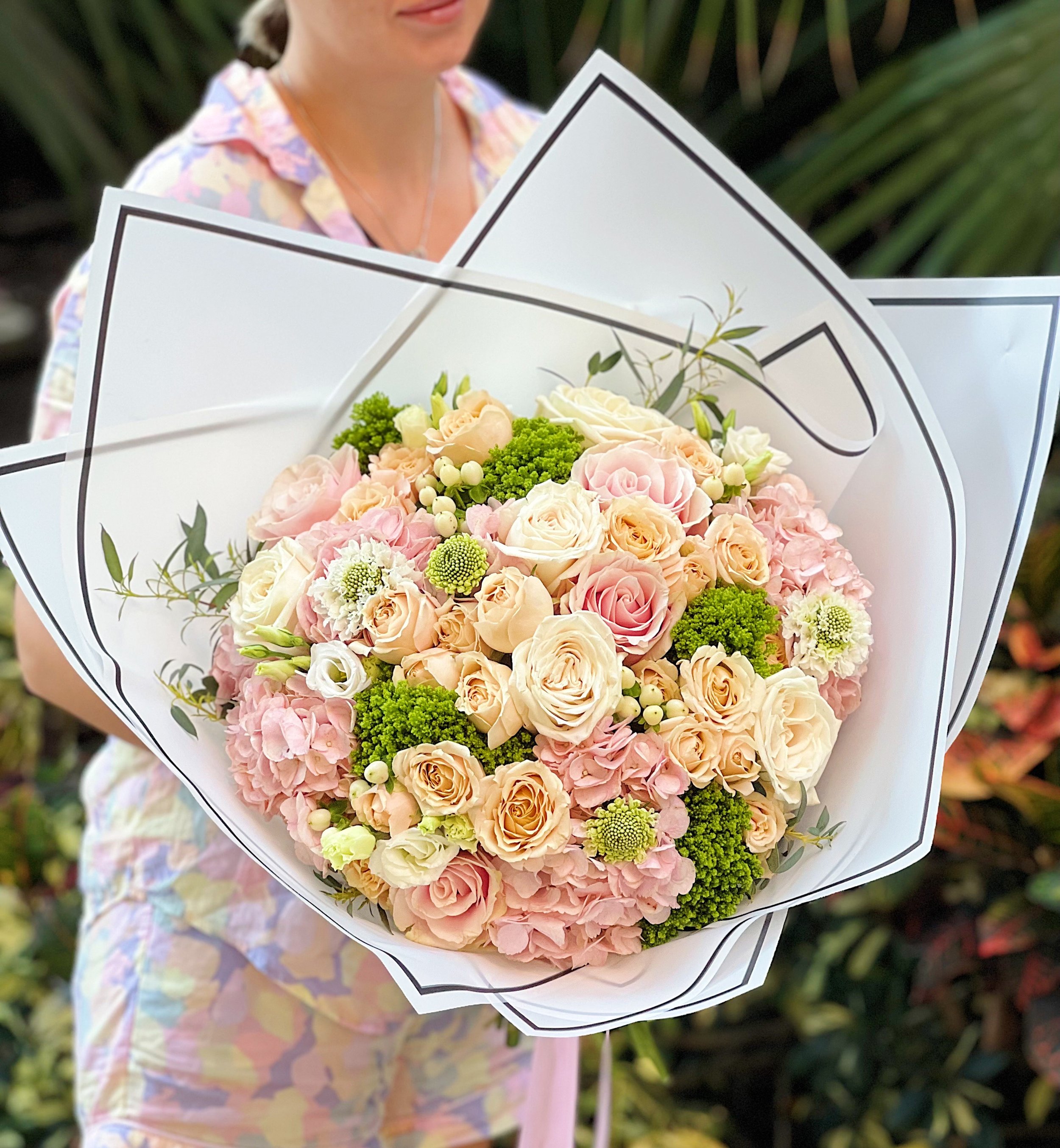A person holding a large bouquet of pink and white roses, complemented by green foliage, wrapped in white paper with black outlines, in an outdoor setting.