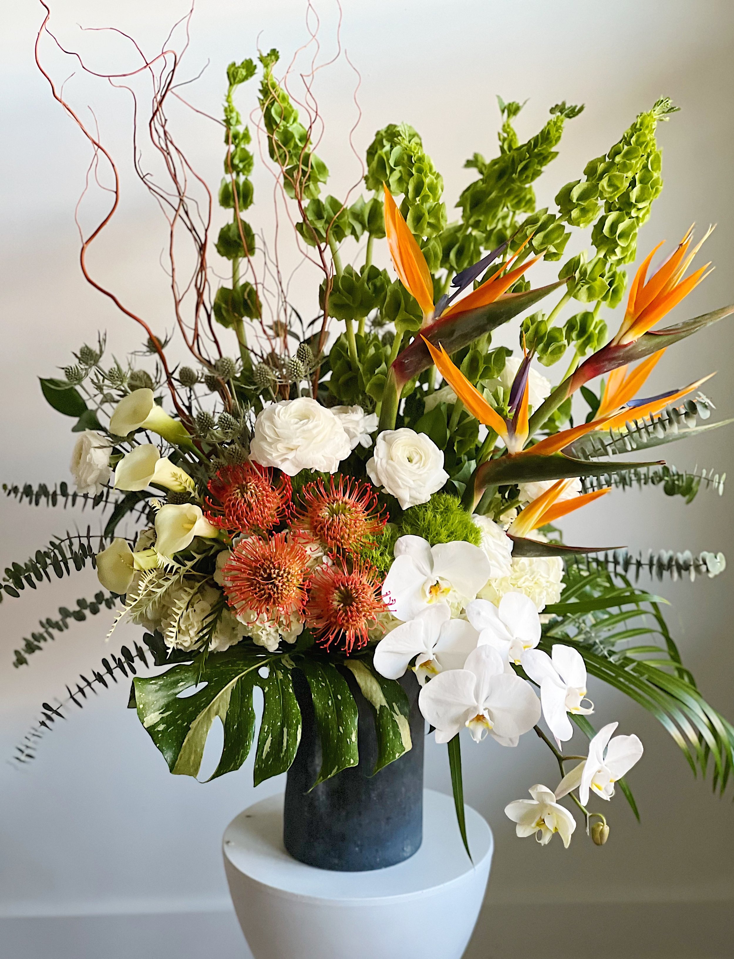 A vibrant tropical flower arrangement in a black vase on a white table, featuring white orchids, orange bird of paradise flowers, red pincushion proteas, white ranunculus, calla lilies, and various greenery.