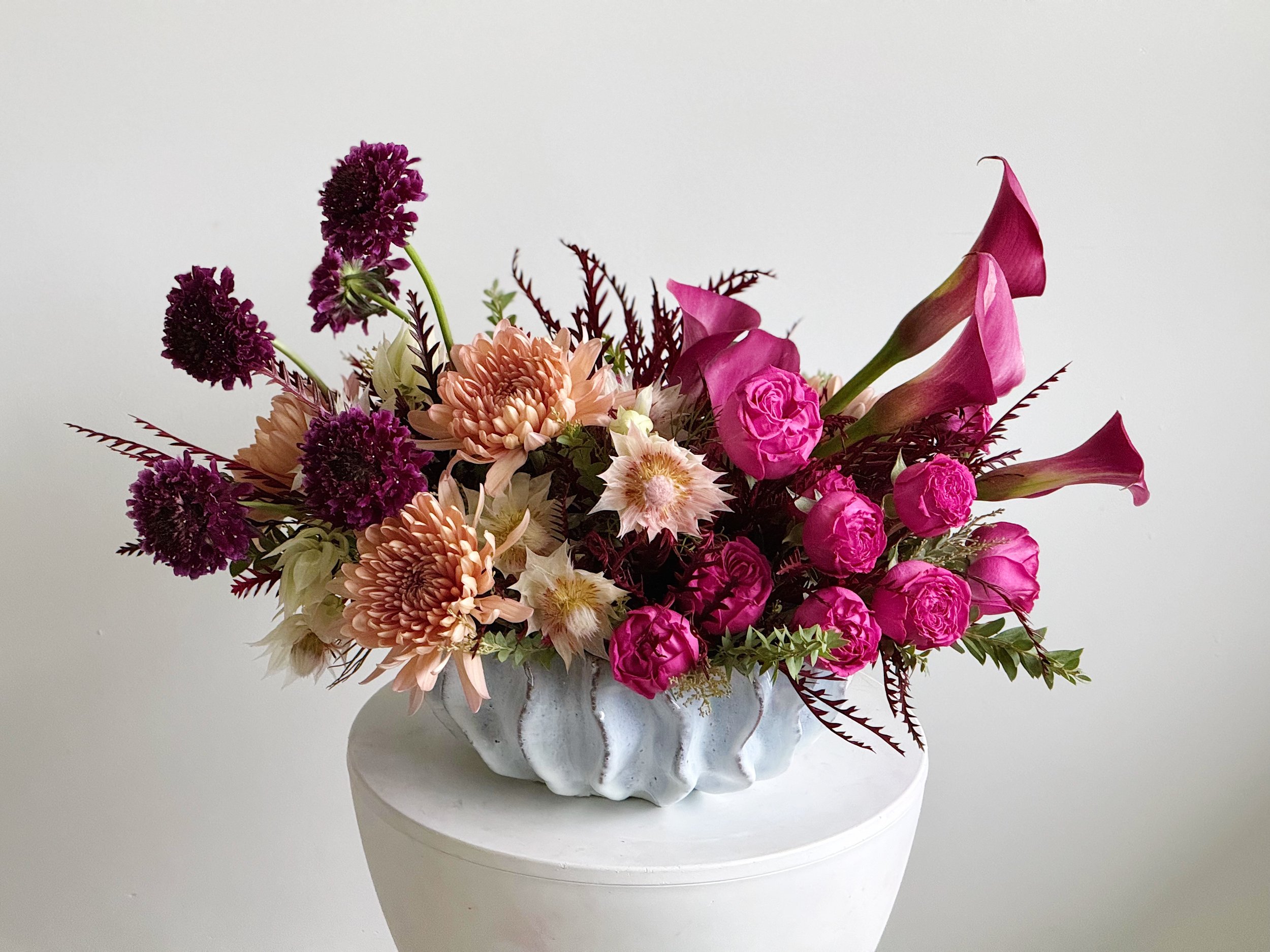 Colorful flower arrangement in a white ceramic vase with pink roses, purple alliums, blush dahlias, and pink calla lilies on a white pedestal against a plain white background.