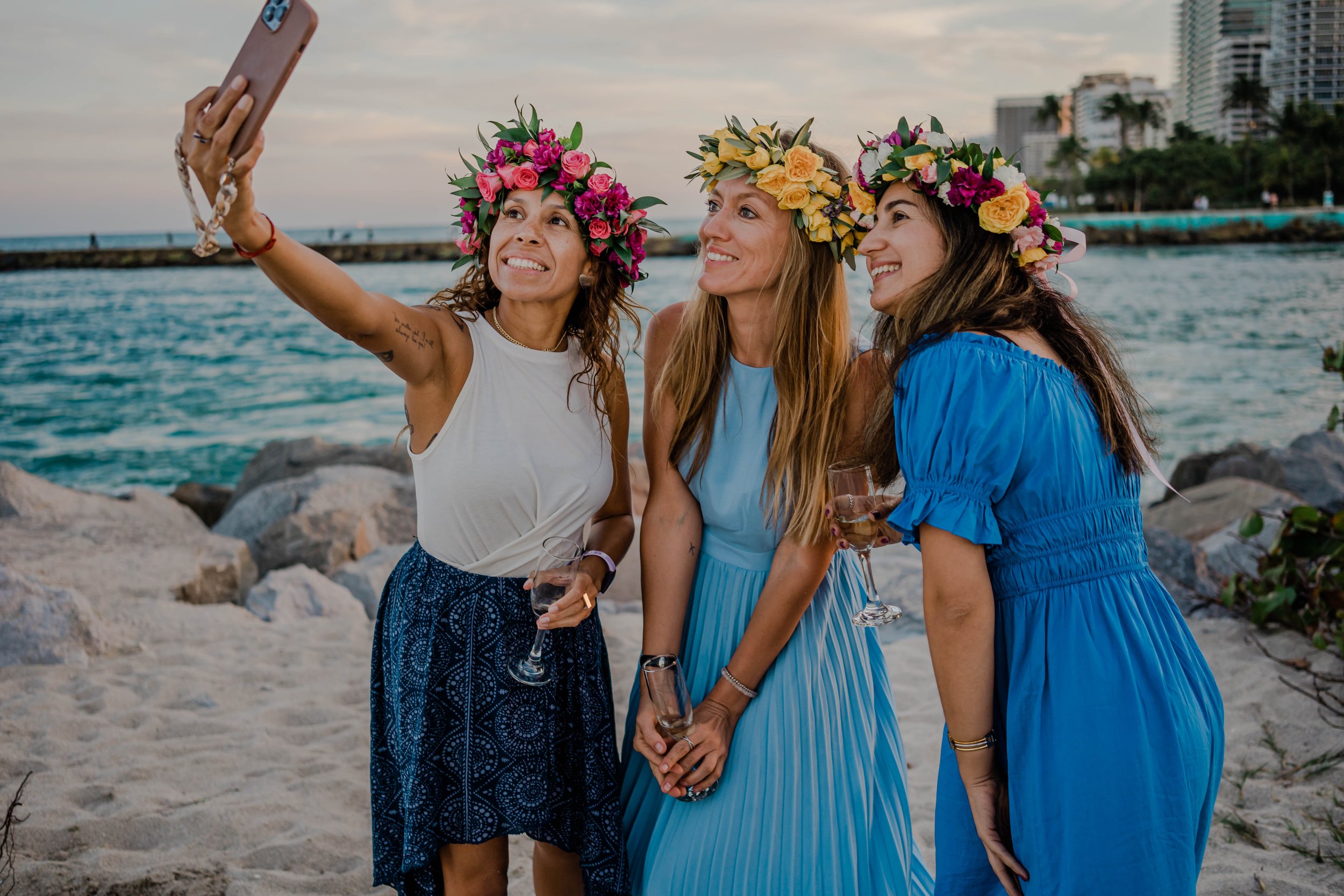 Three women wearing flower crowns taking a selfie on a beach during daytime, holding wine glasses with buildings and the ocean in the background.