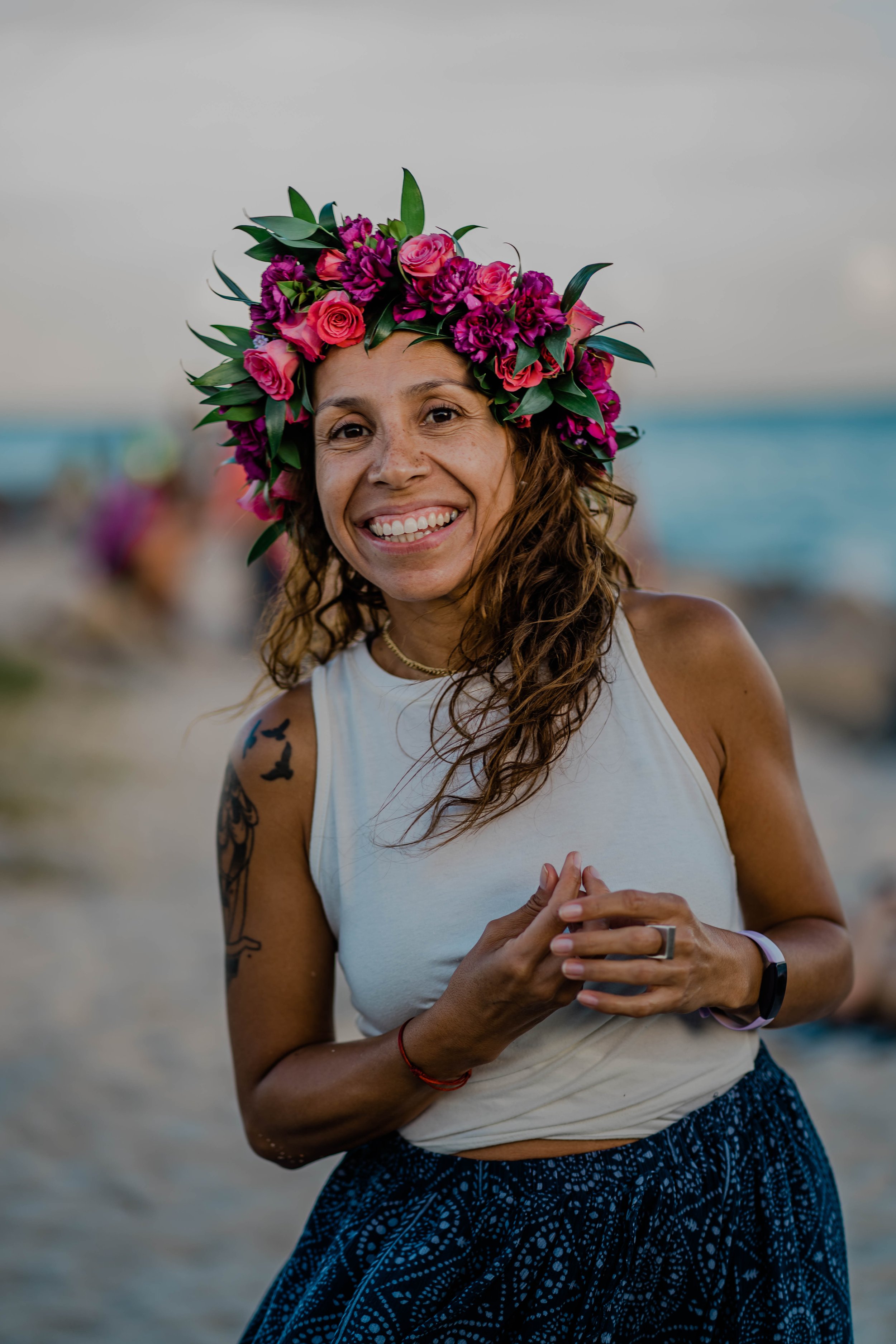 A woman smiling at the beach wears a white sleeveless top, blue patterned pants, a crown of pink and purple flowers, and has a tattoo of birds on her arm.