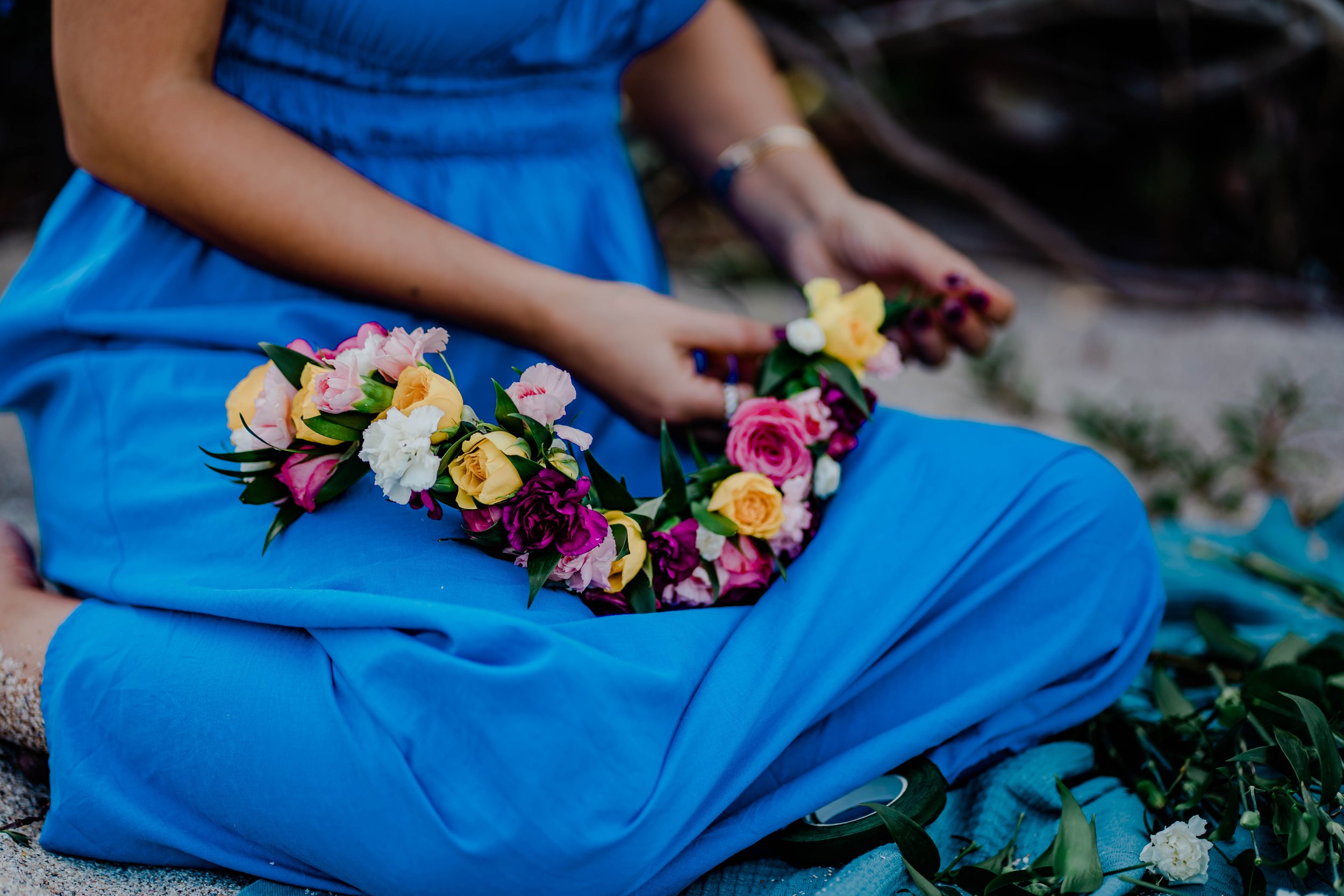 Person dressed in a blue outfit sitting on the ground, holding a floral headpiece made of pink, yellow, white, and purple flowers with green leaves.