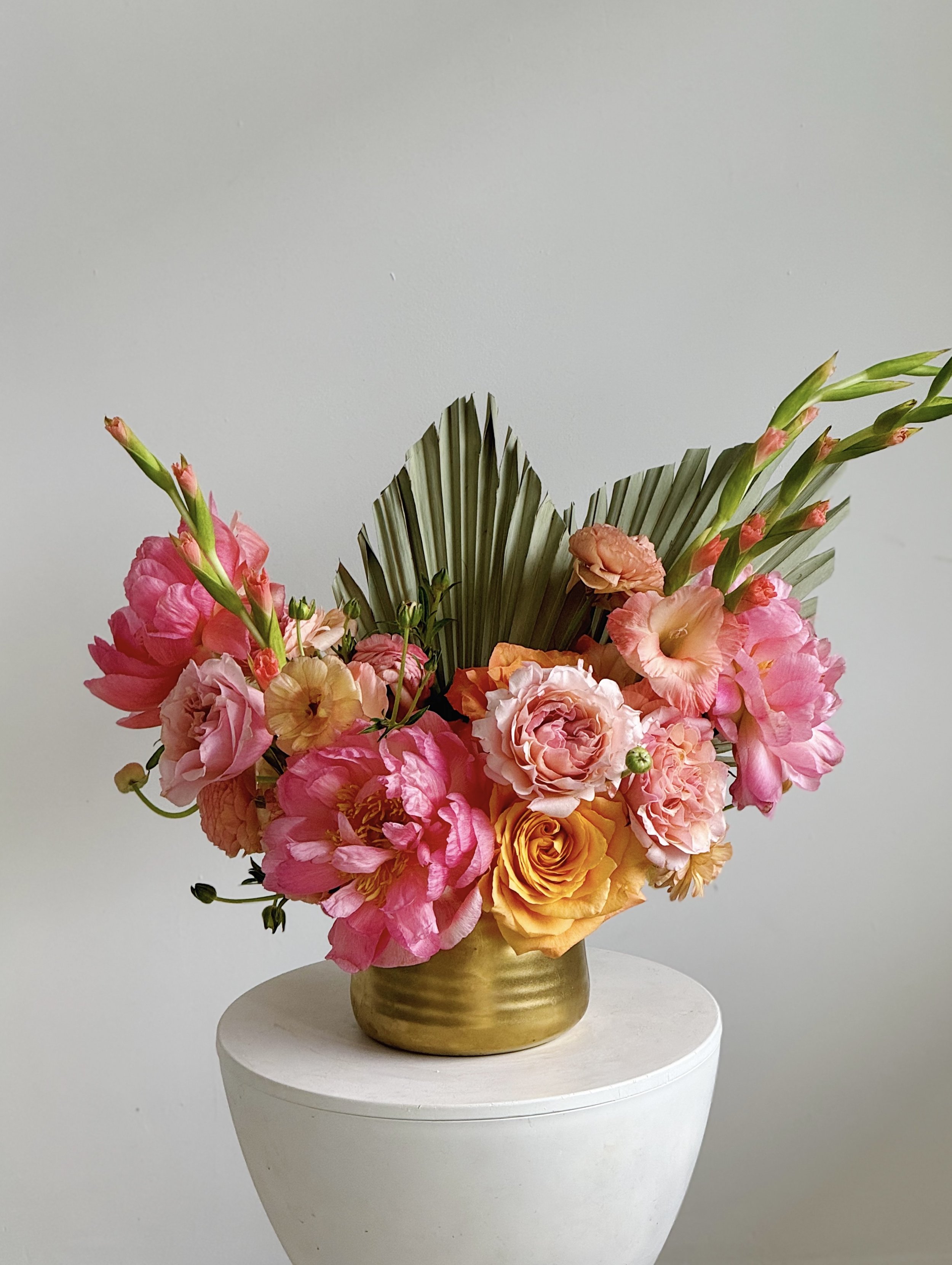A colorful bouquet of pink, peach, and yellow flowers in a greenish-gold vase on a white pedestal against a plain background.