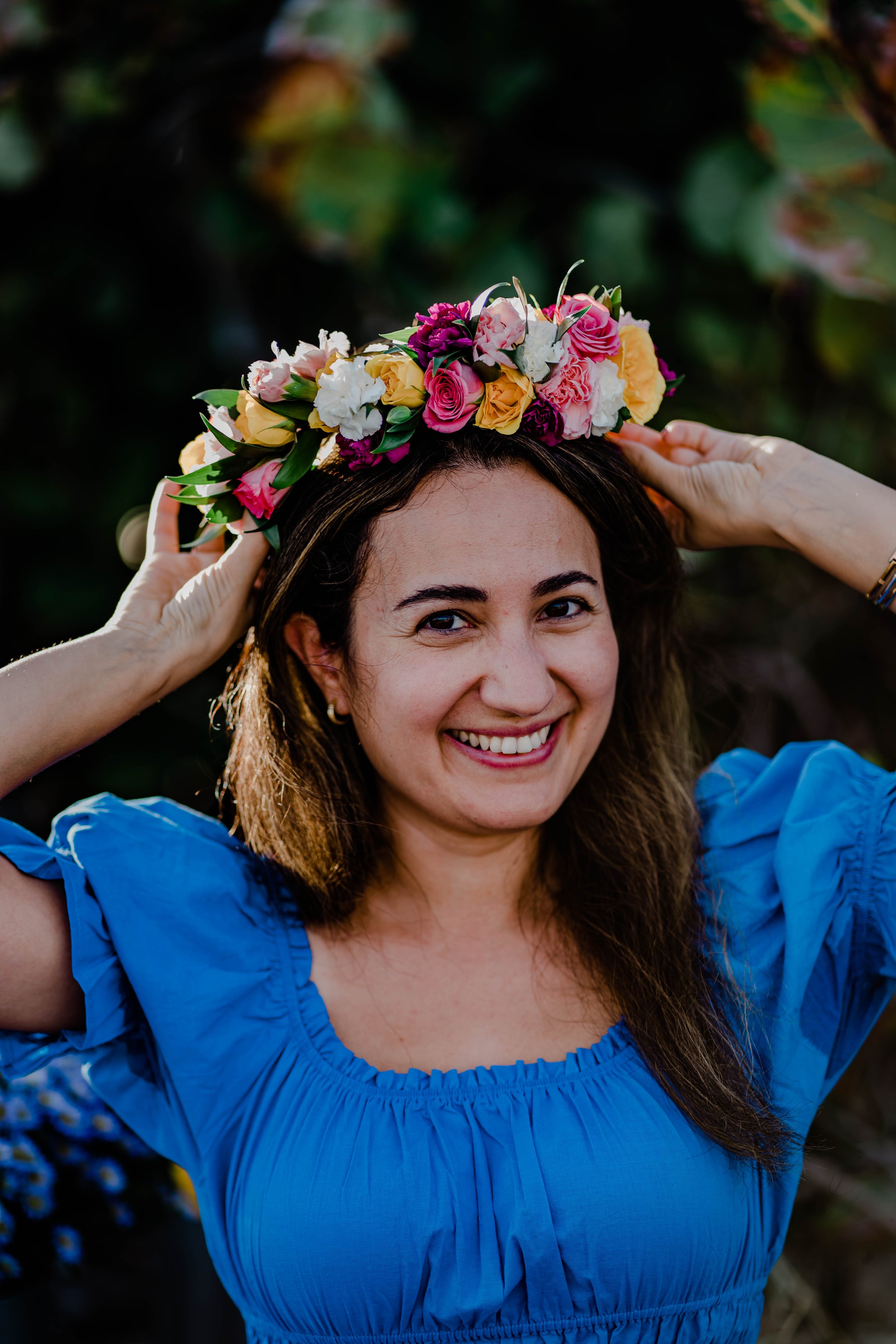 Woman wearing a blue dress placing a colorful flower crown on her head outdoors, smiling at the camera.