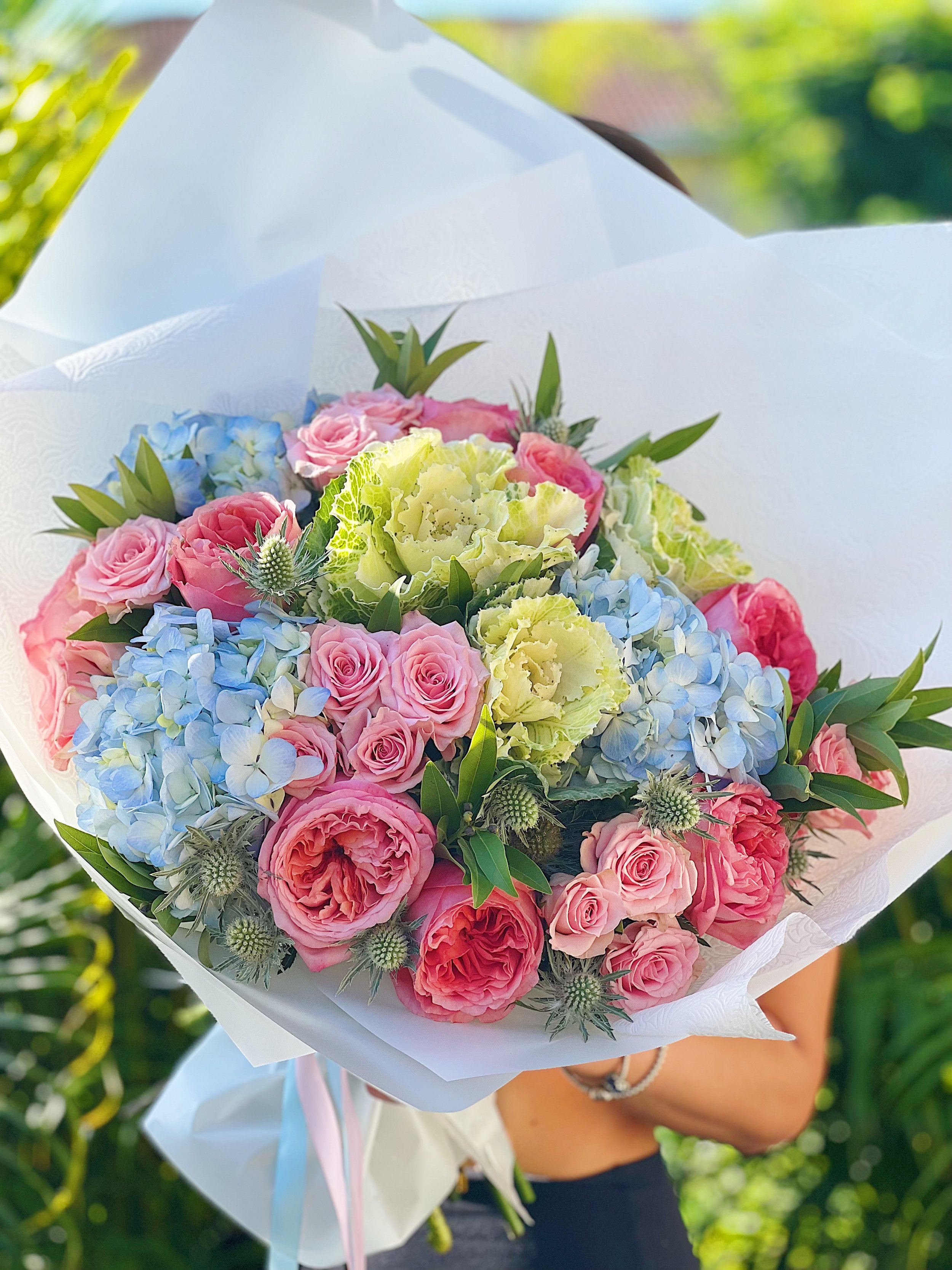 A person holding a large bouquet of mixed flowers including pink roses, blue hydrangeas, yellow ornamental kale, and other greenery, wrapped in white paper with pastel ribbons, outdoors with greenery in the background.