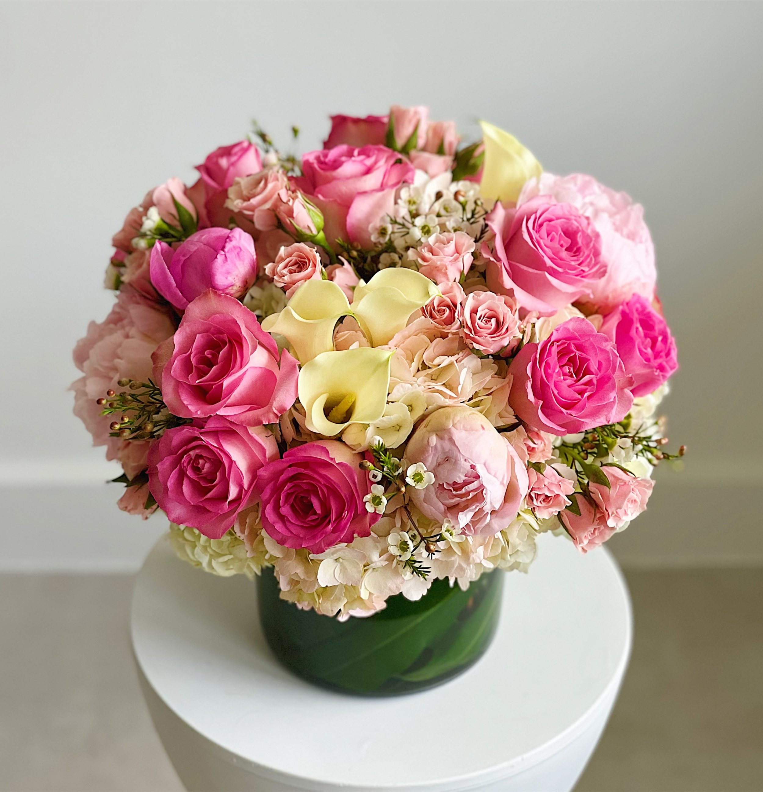 A round bouquet of pink and white roses, calla lilies, and small filler flowers in a green glass vase on a white round table.