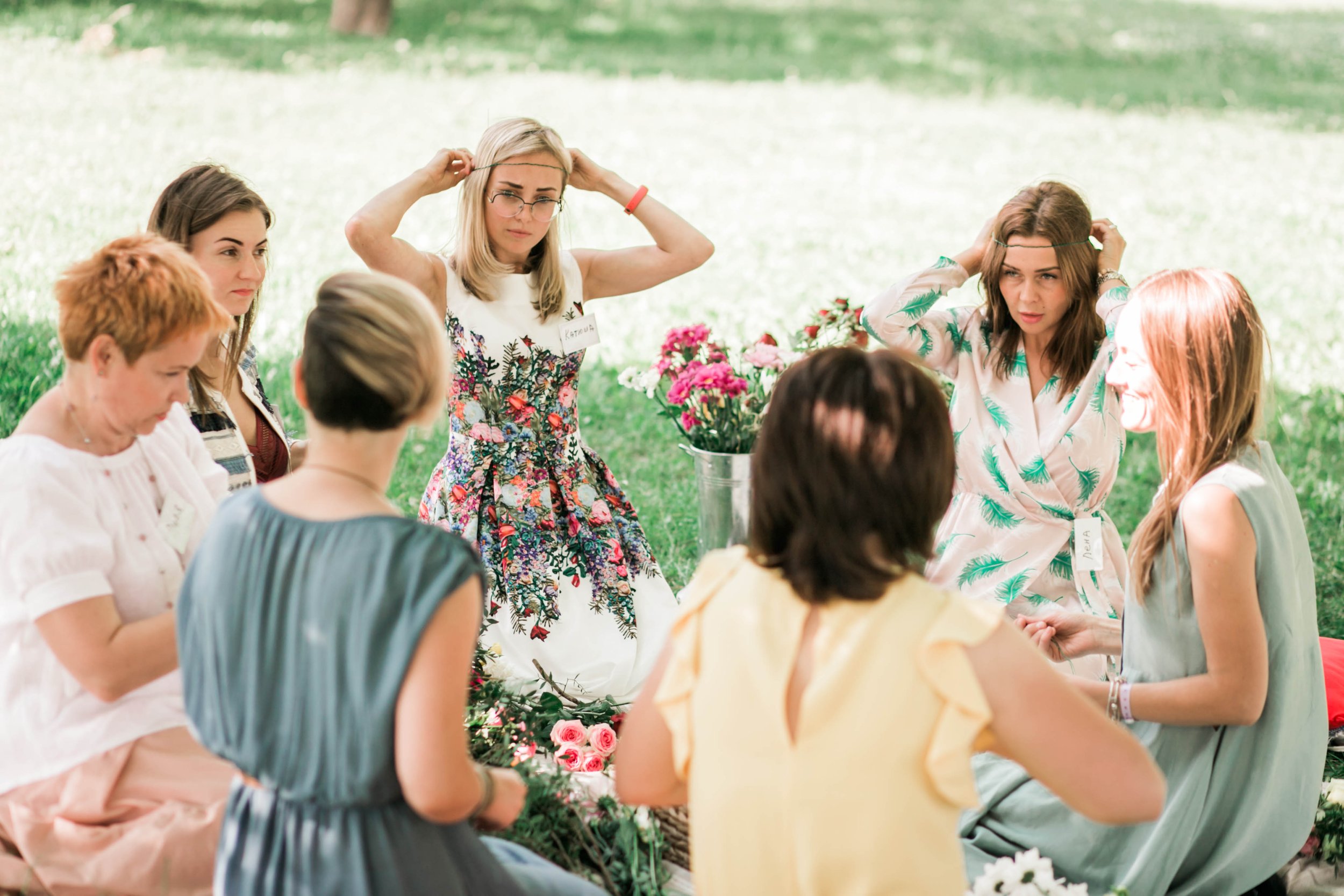 Group of women sitting on grass in a circle during a flower arranging workshop outdoors, with flowers and greenery surrounding them.