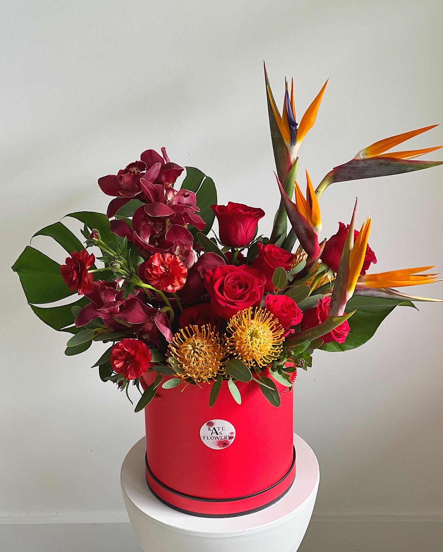 A vibrant bouquet of red roses, pink orchids, orange bird of paradise flowers, and yellow pincushion proteas in a red round box with a "Kate's Flowers" sticker, placed on a white pedestal against a plain background.
