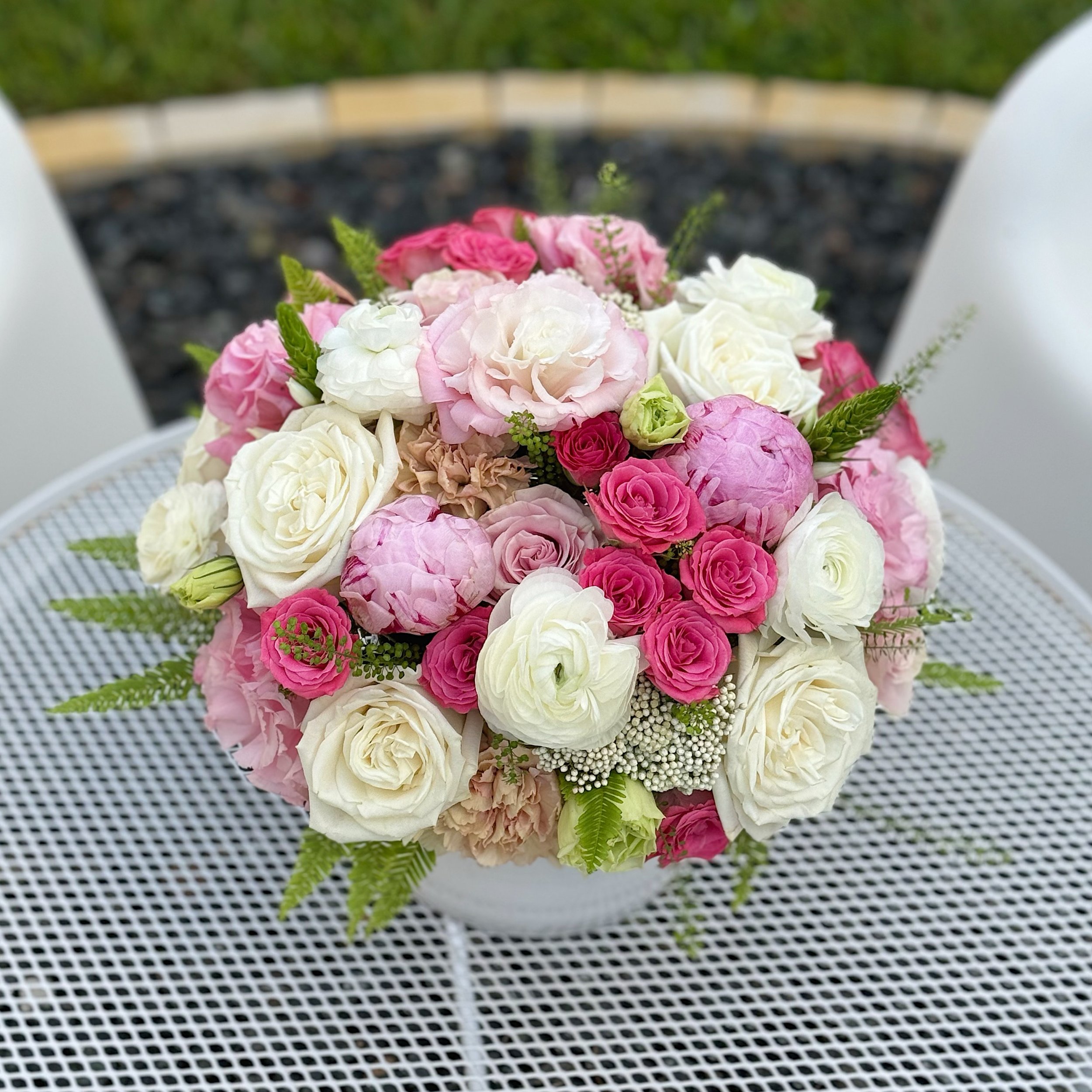 A colorful bouquet of pink, white, and cream roses and other flowers arranged on a white metal table outside.