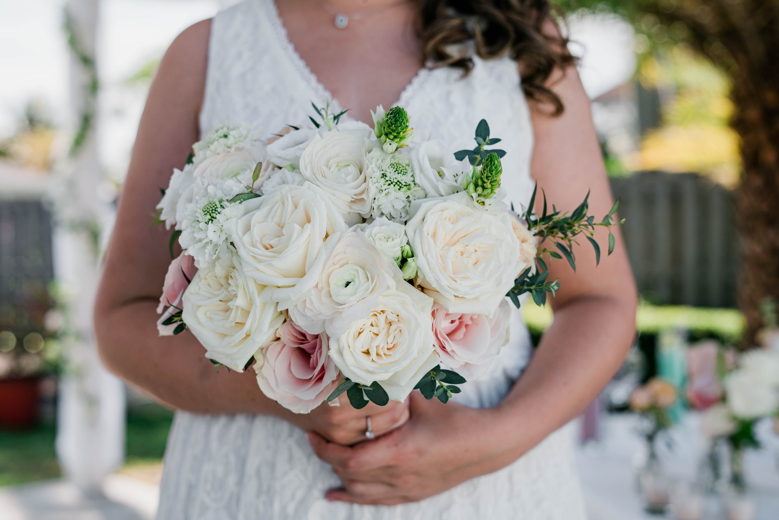 A woman in a white sleeveless dress holding a bridal bouquet of white and pale pink roses and other white flowers, outdoors with blurred background.