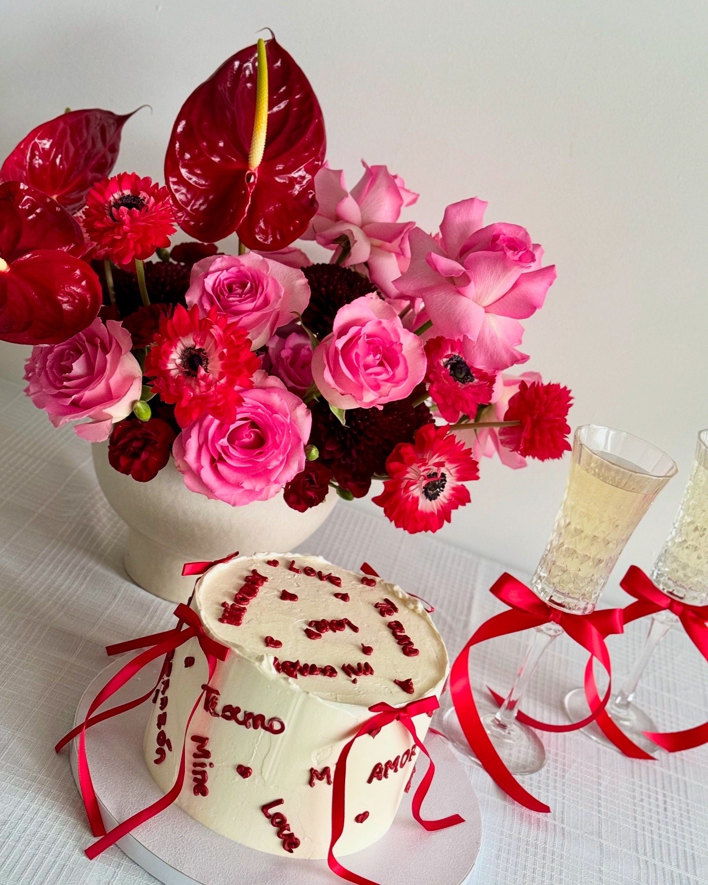 A white heart-shaped cake decorated with red ribbon and love words in Spanish, topped with small red hearts. Next to the cake are two champagne glasses with red ribbons tied around their stems. Behind them, a large bouquet of pink and red flowers, in