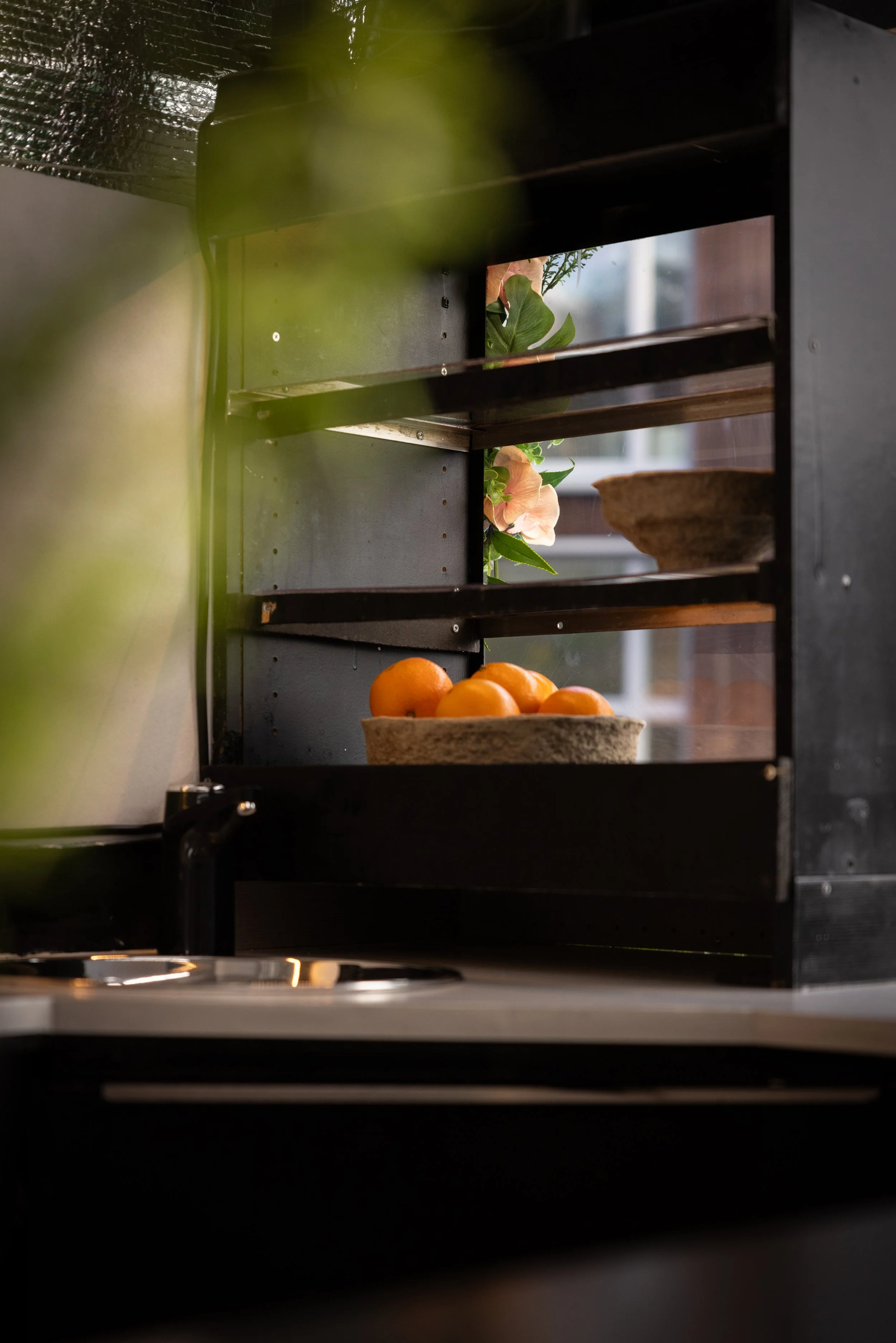A black kitchen shelf holding a bowl of oranges, pink flowers, and a small plant, with a window in the background.