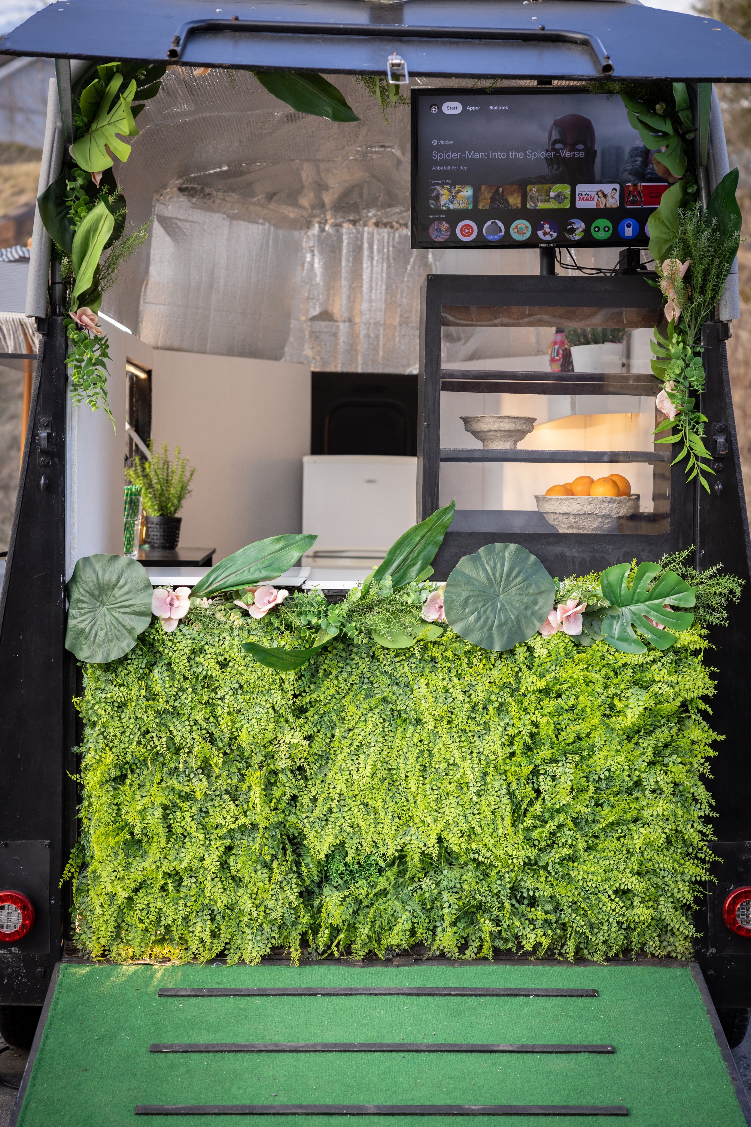 A mobile food or beverage cart decorated with green plants and pink flowers, featuring a flat-screen TV, a small counter with a bowl of oranges, and a lush green foliage backdrop.