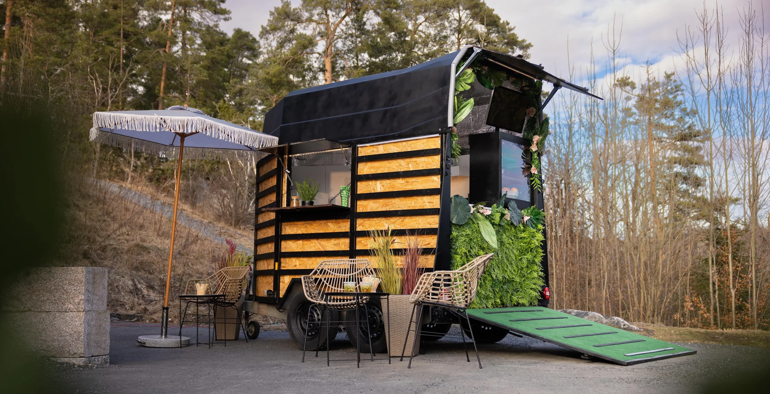 A food truck decorated with green plants and flowers, with an outdoor seating area including a table, chairs, and a white umbrella, set against a backdrop of trees.