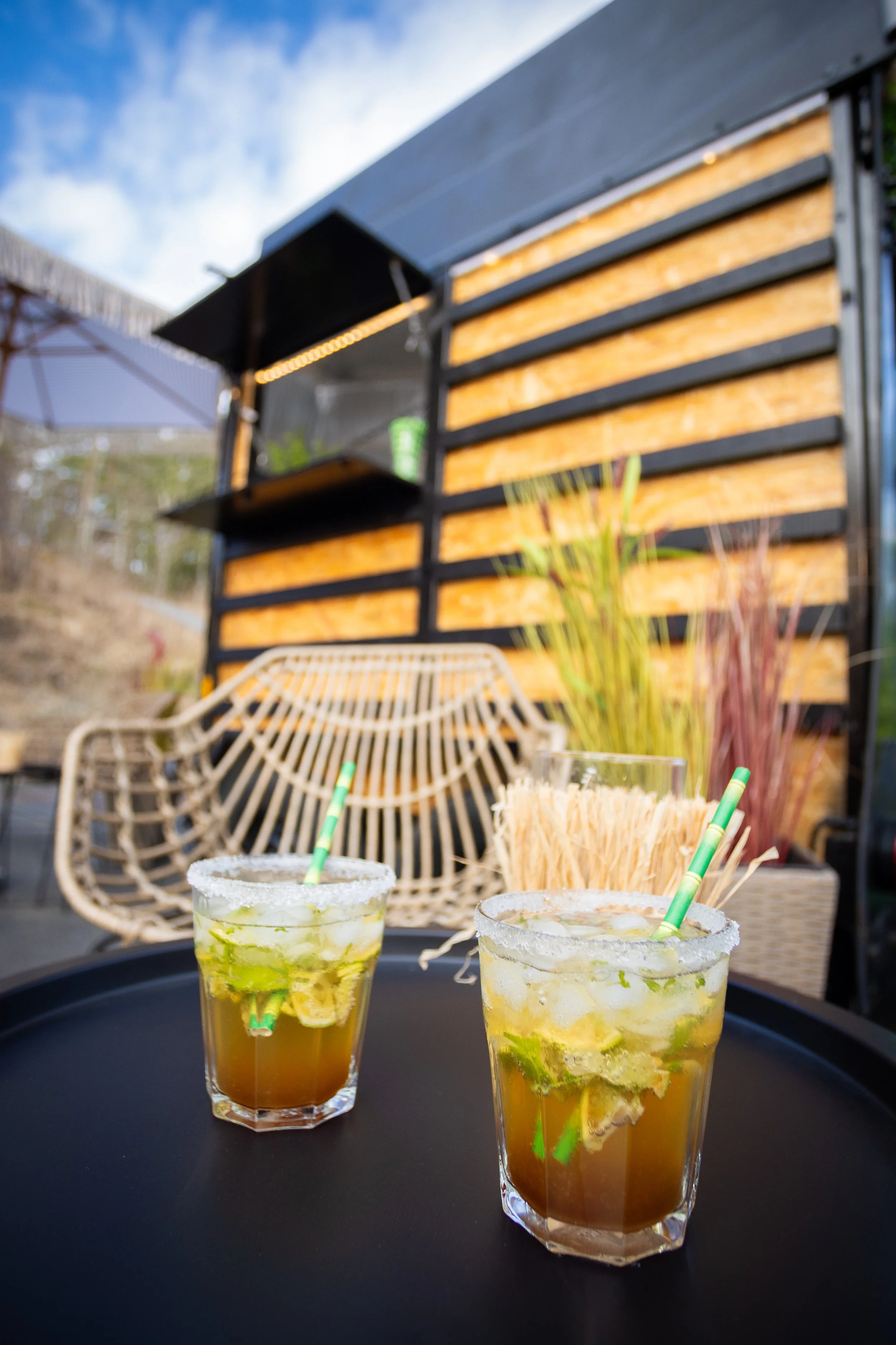 Two margarita cocktails with lime slices on the rim, on a black tray outdoors. In the background, a rattan chair, tall plants, and a wooden and black metal structure.