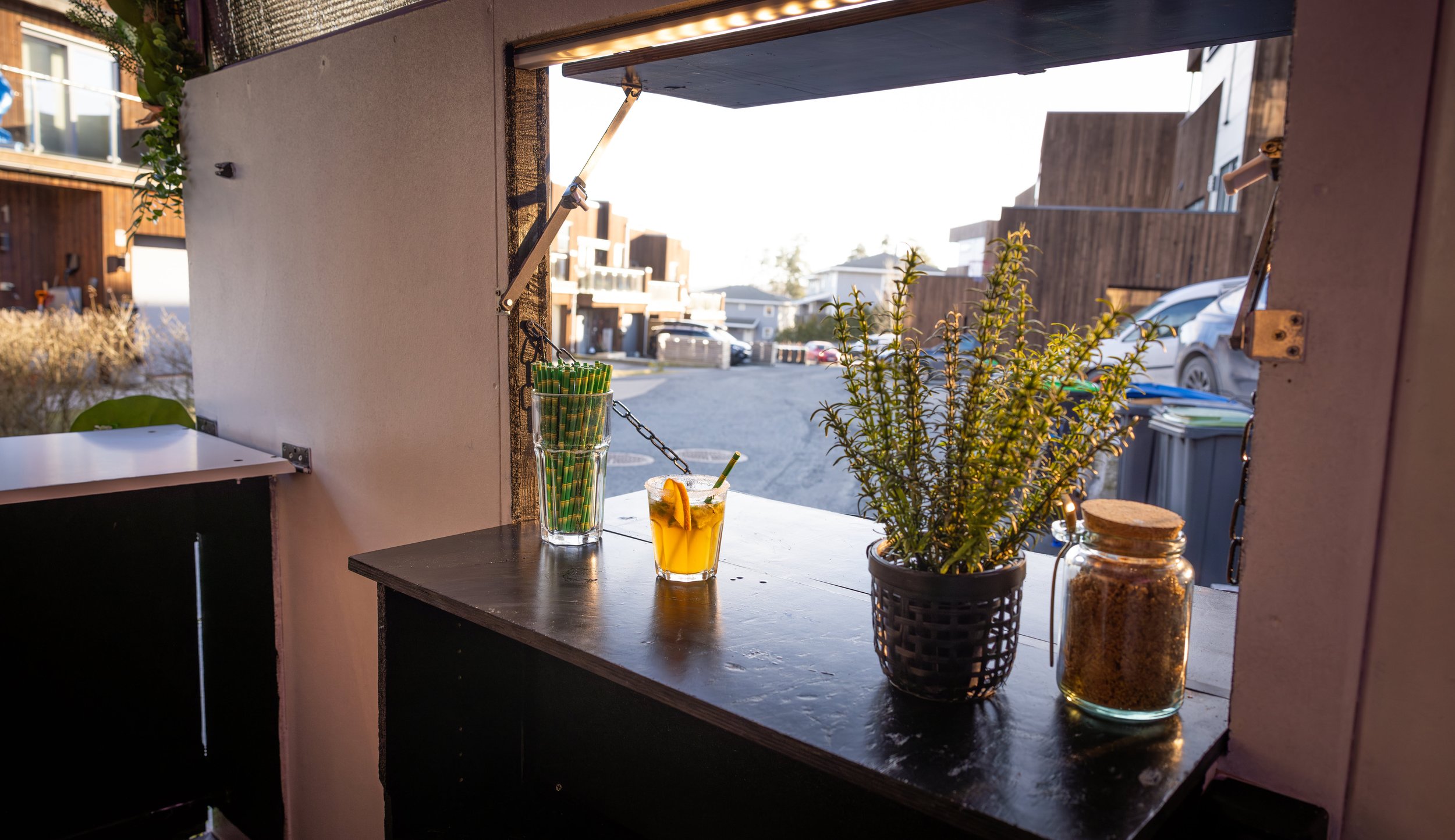 Inside a cafe or restaurant with a large window/door opening to the outside. On the windowsill, there are two drinks: a tall glass with green and white striped straws, and a shorter glass with a yellow drink garnished with fruit. There is also a pott