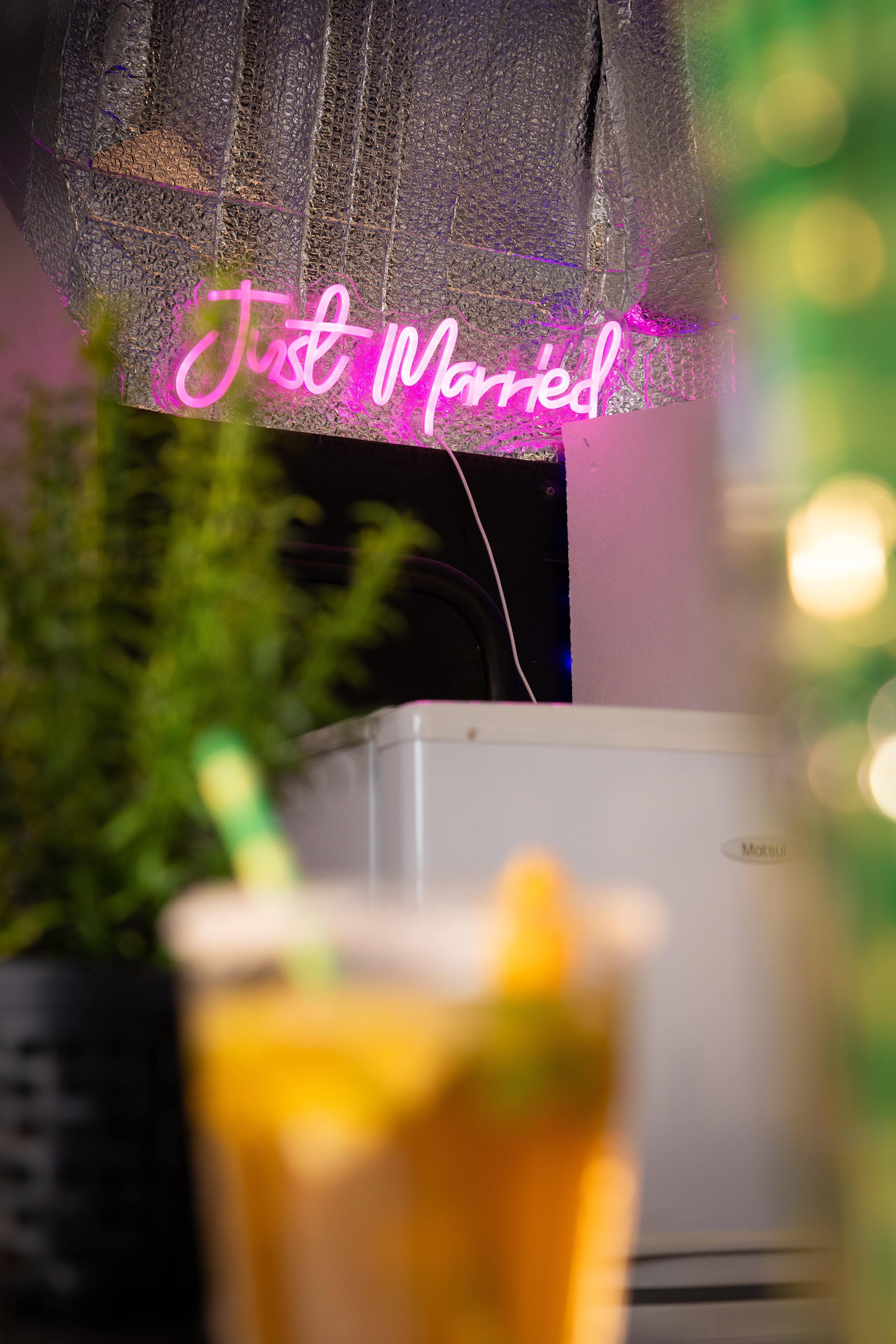 A neon pink sign that says "Just Married" is mounted on a textured metallic wall in the background, with some fuzzy elements of greenery and a drink with a straw in the foreground.