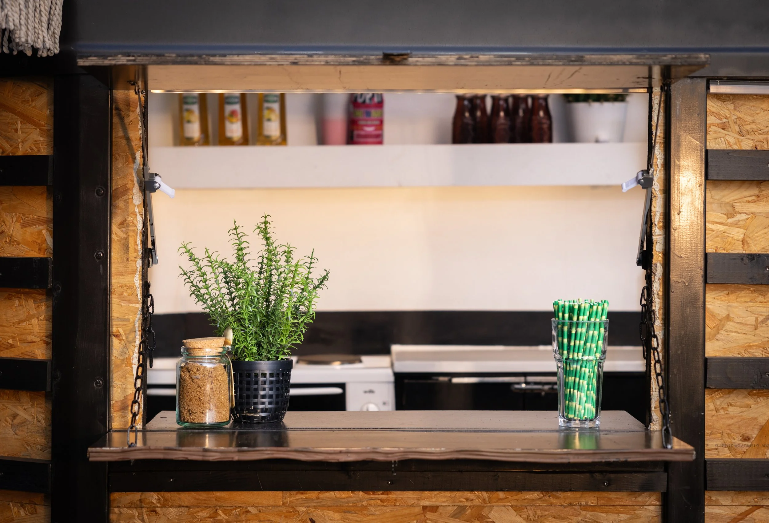A wooden shelf with a potted plant, a jar of brown sugar, a glass of green and yellow striped straws, and a black container, with a kitchen in the background.