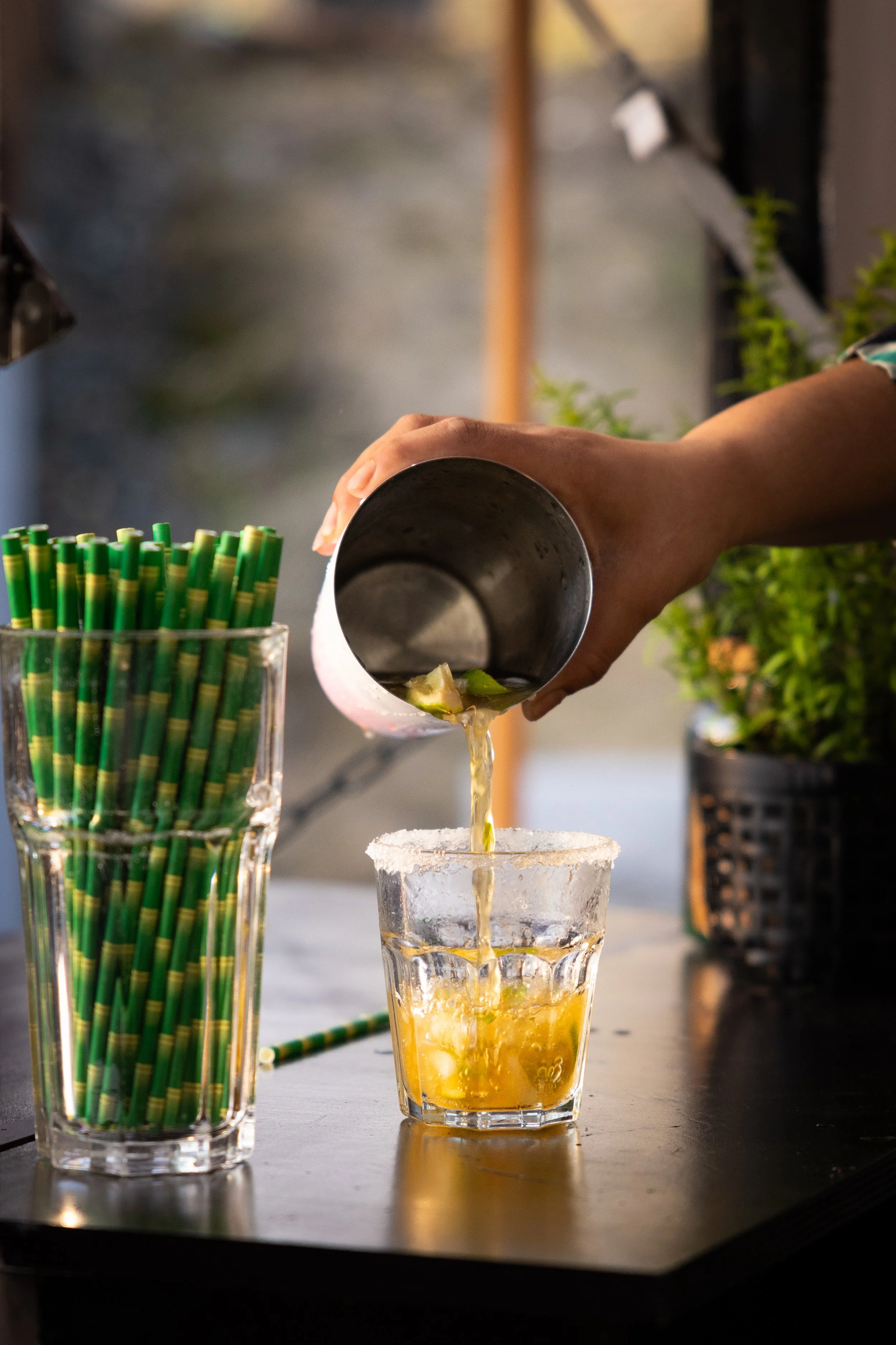 Person pouring a yellow beverage, possibly juice, into a glass rimmed with salt, with a container of green and yellow straws nearby.
