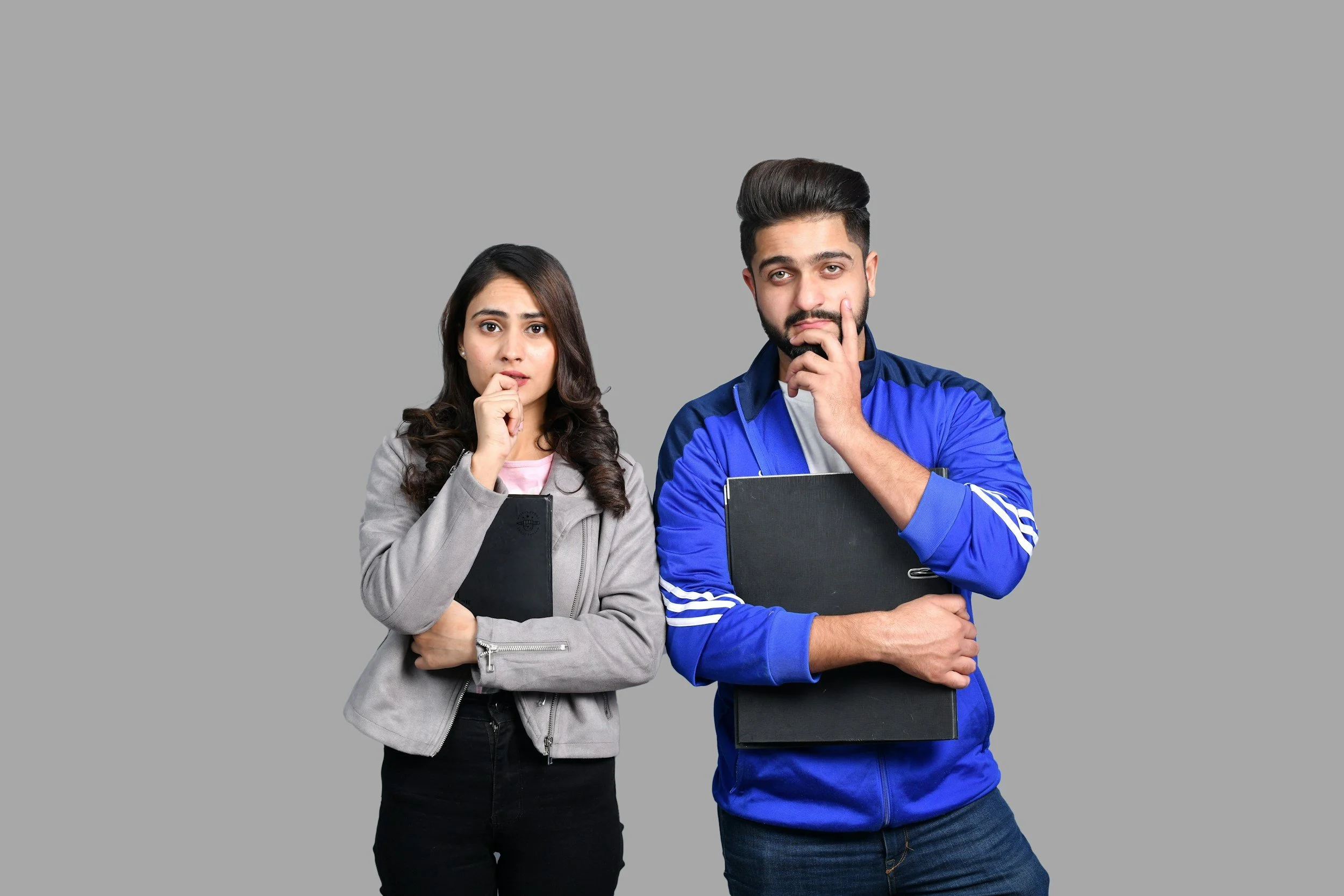 A young woman and a young man standing side by side against a gray background, both holding black folders and appearing contemplative.