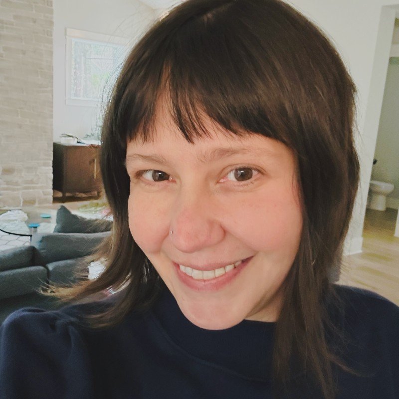 A woman with short brown hair, smiling in a living room with a fireplace and a couch in the background.