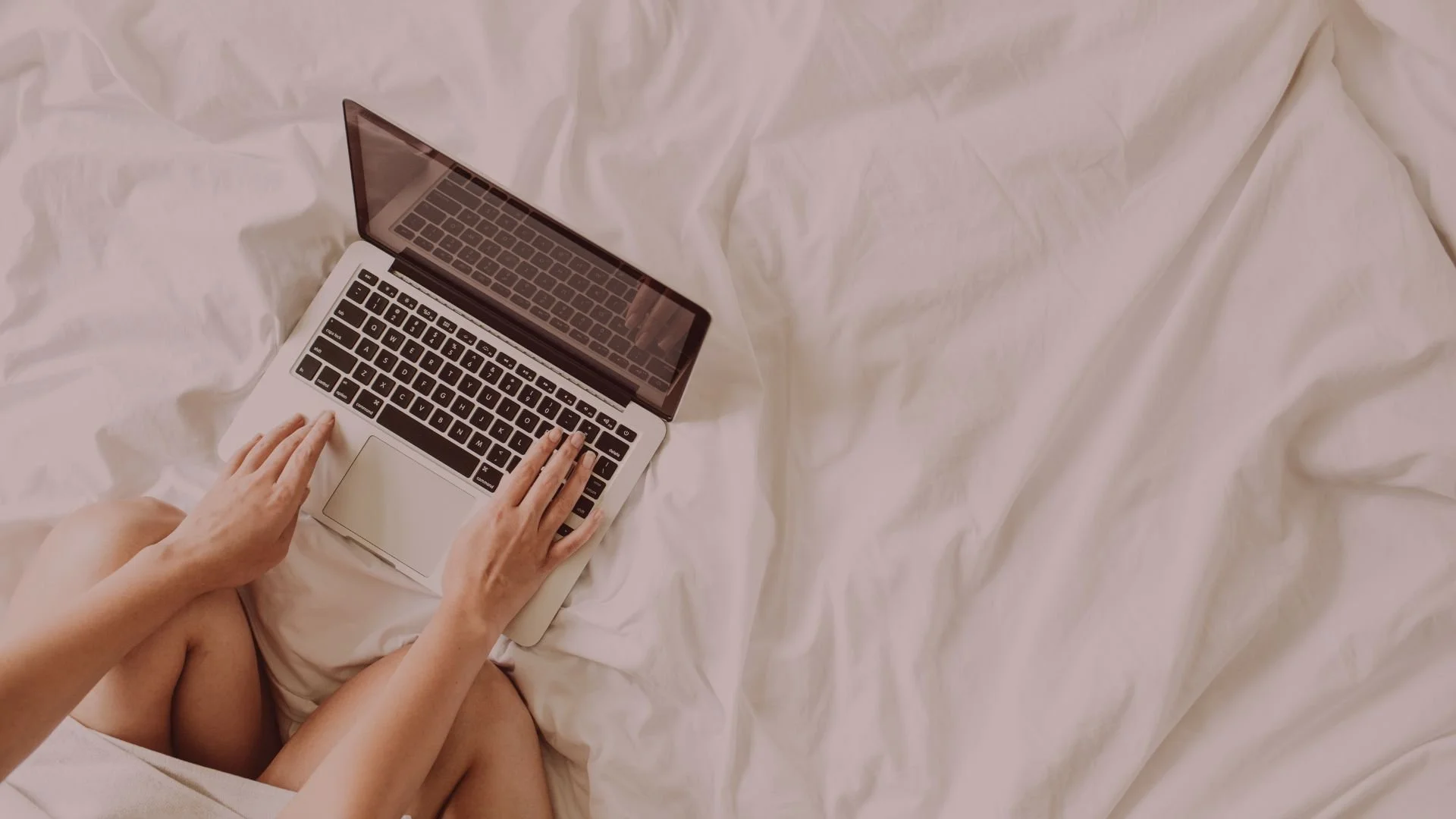 Person sitting on a bed using a silver MacBook laptop with a black keyboard, surrounded by white bedsheets.
