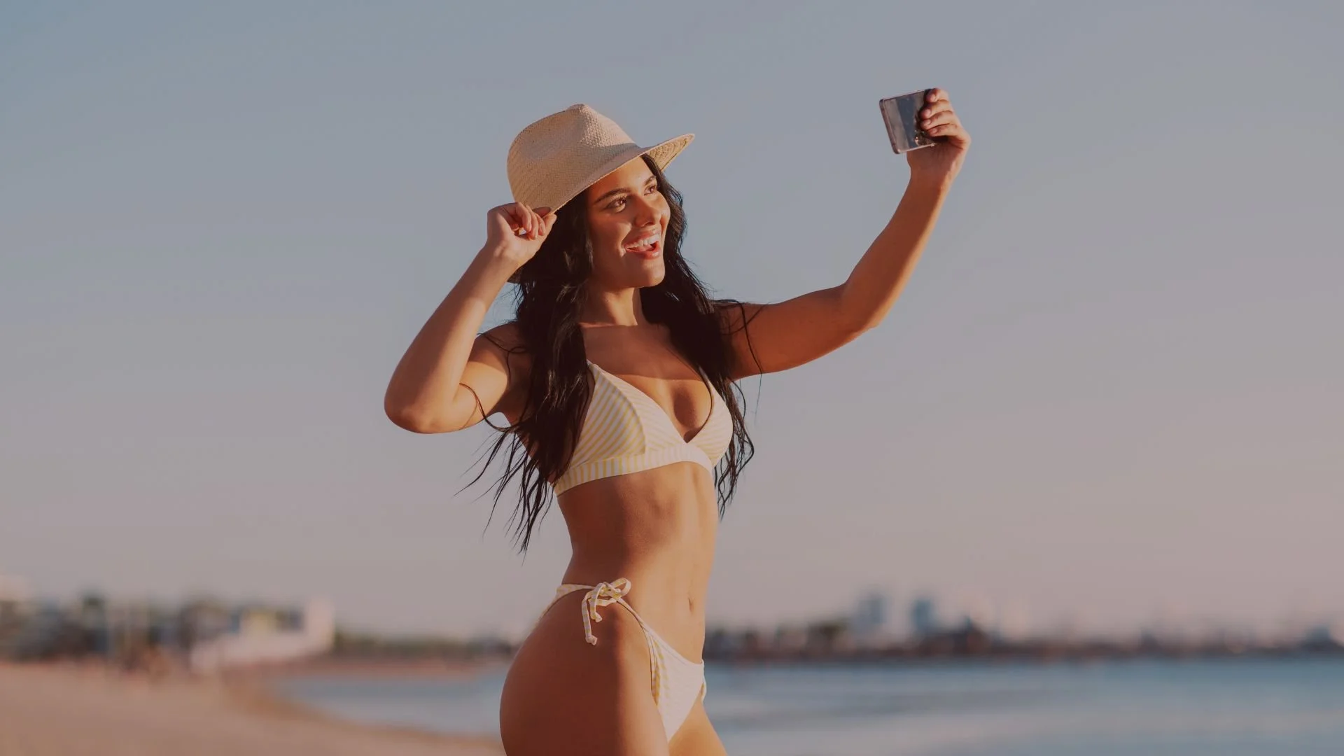 Woman in a bikini taking a selfie on the beach wearing a sun hat.