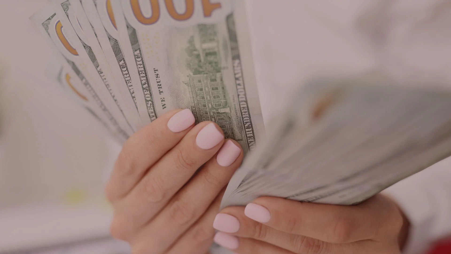 Close-up of a hand with light pink nail polish holding a fan of hundred-dollar bills.